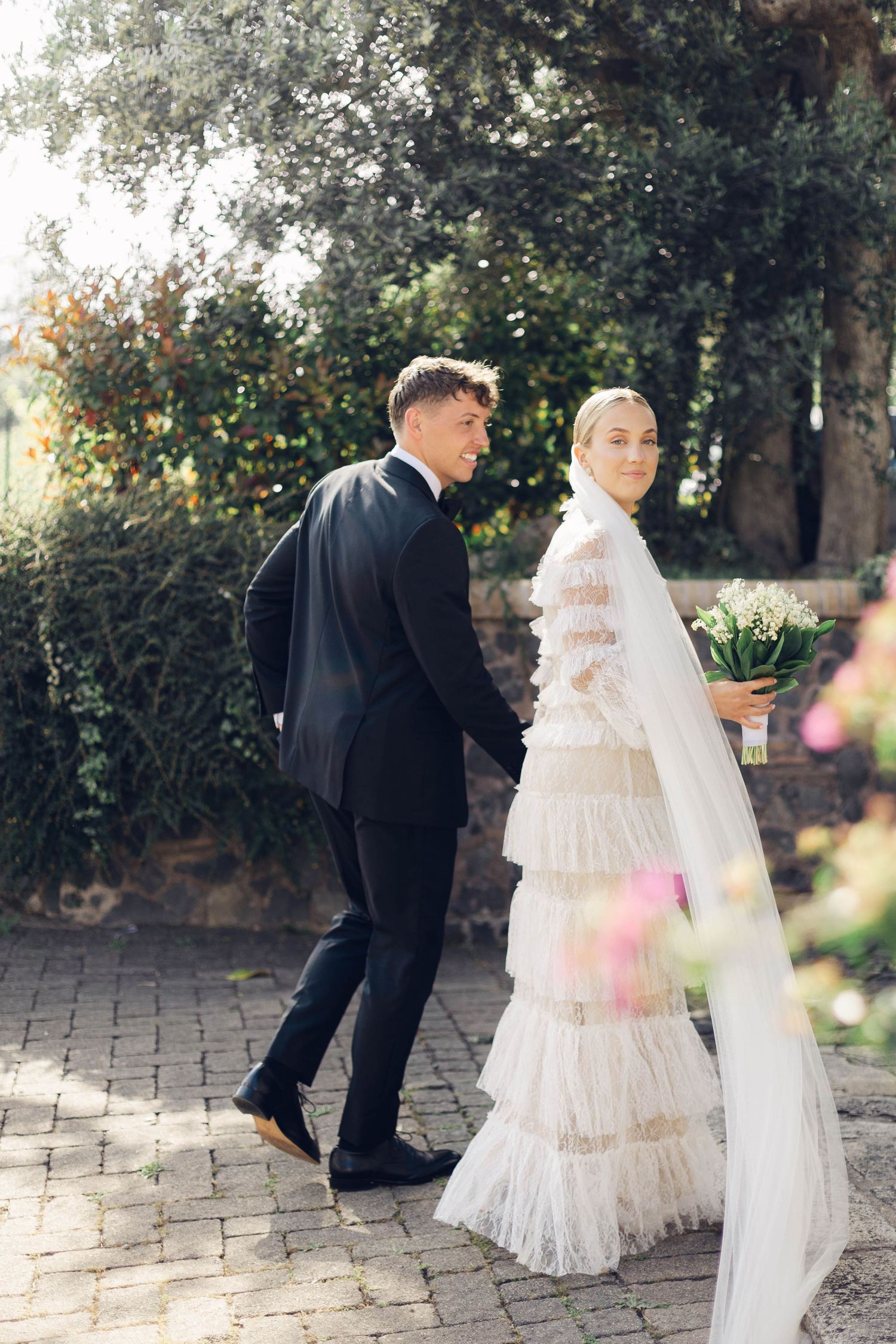 A couple in wedding attire walk along a garden path with sunlit trees in the background.