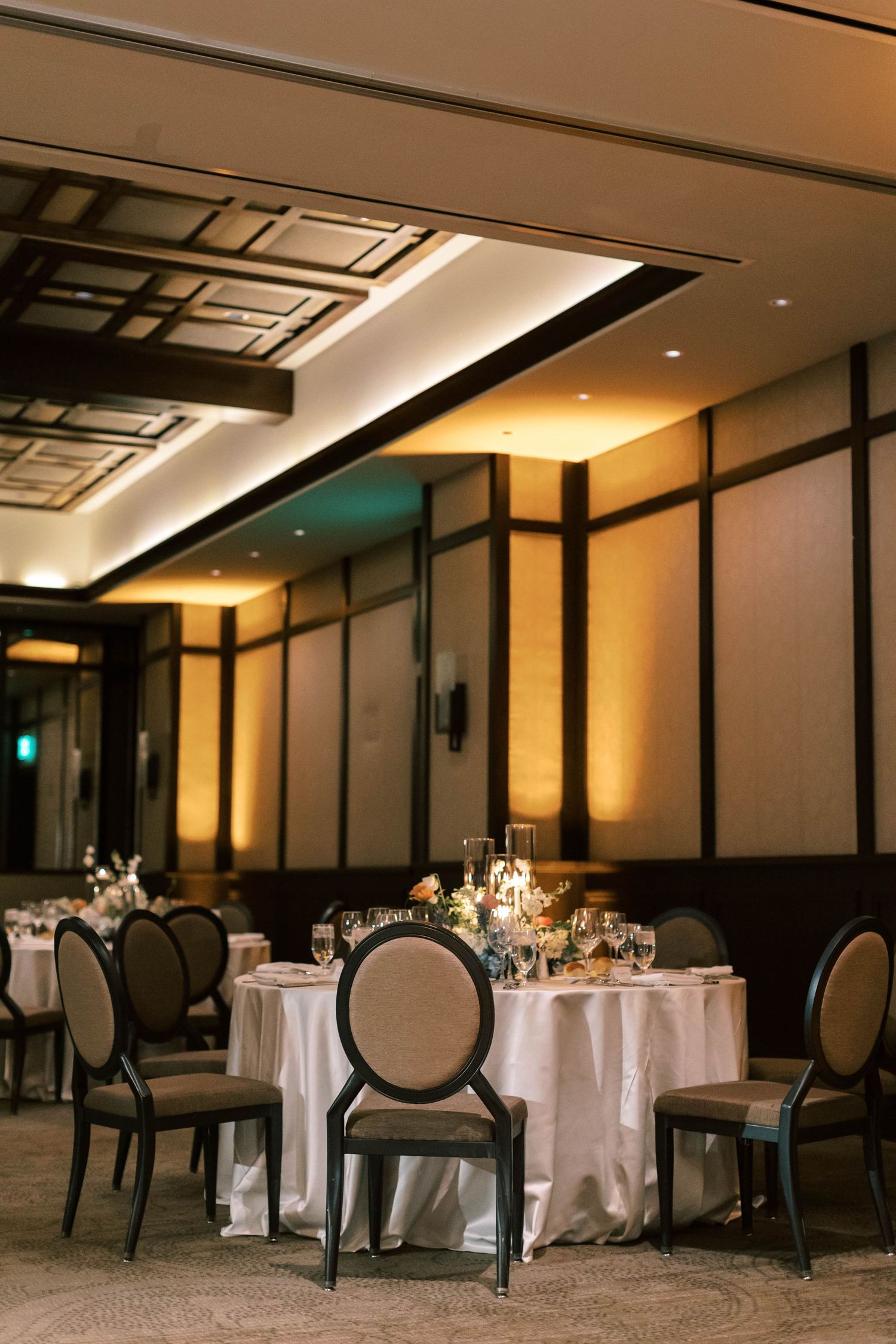 Elegant wedding reception venue with round tables, white linens, and modern oval-back chairs in a warmly lit ballroom.