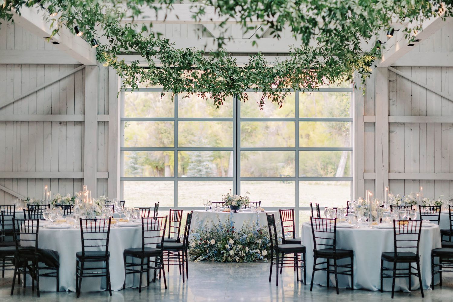 Modern barn reception setup with white tables, black chairs and hanging greenery decor.