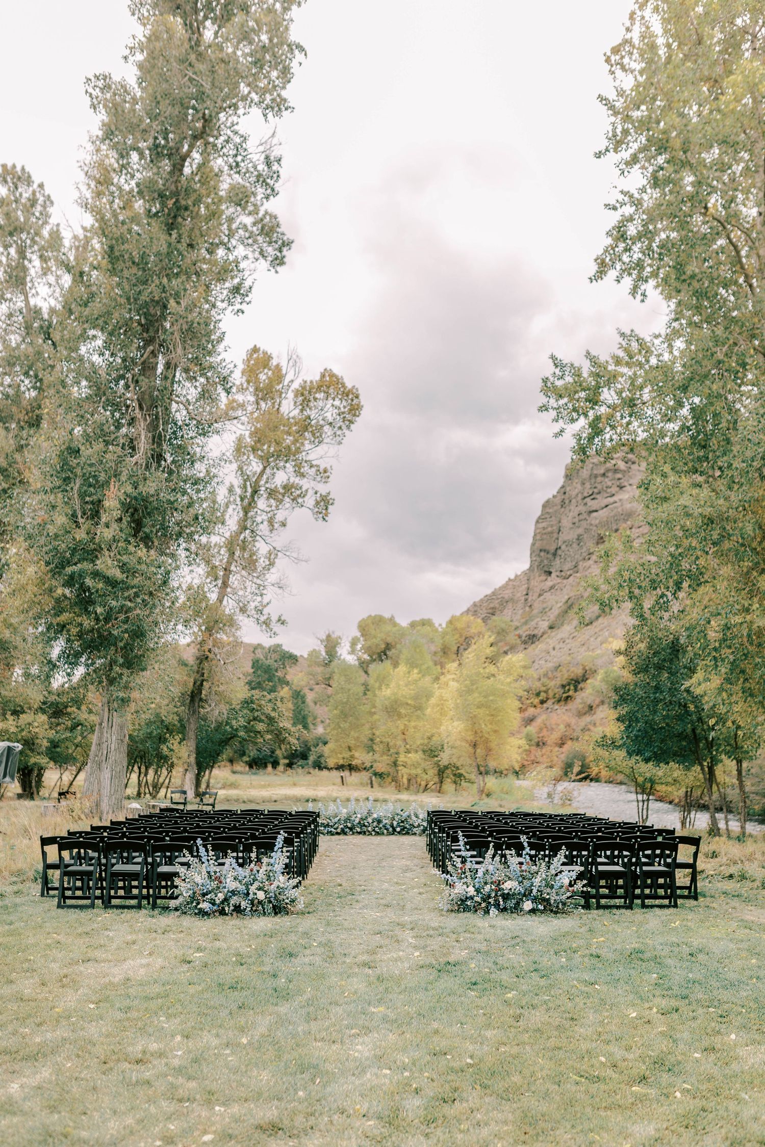 Outdoor wedding ceremony setup with black chairs arranged in rows, framed by tall trees and rocky cliffs.