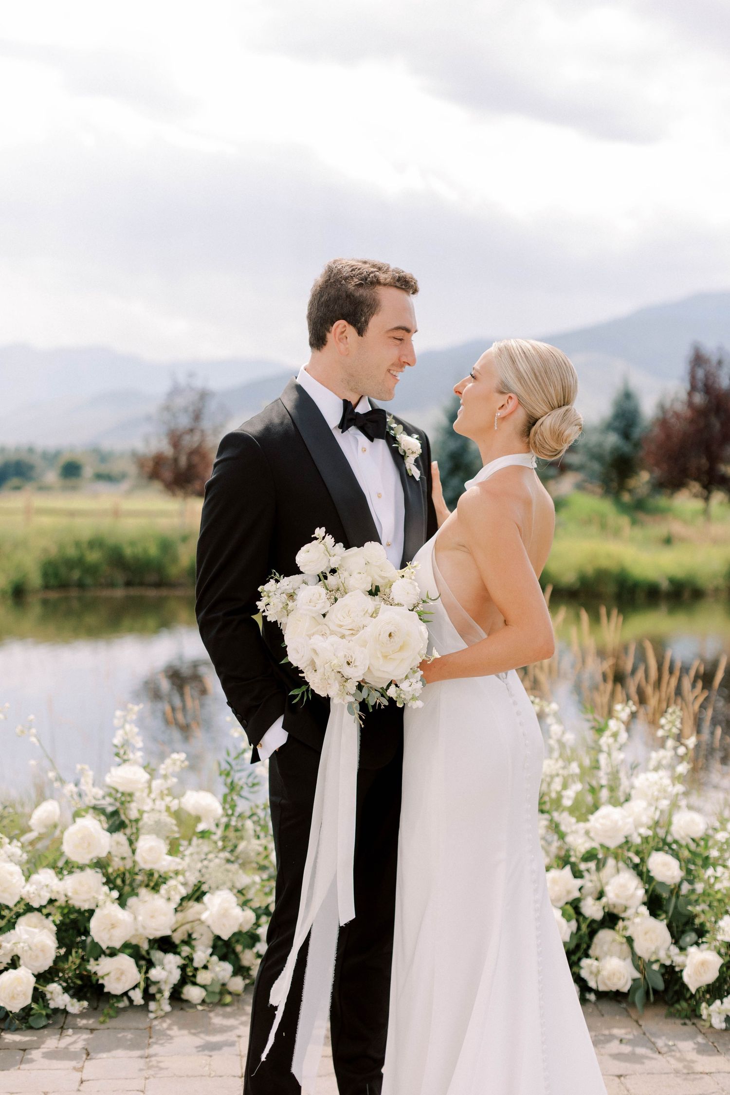 Wedding couple stands by pond with mountain view and white floral arrangements.