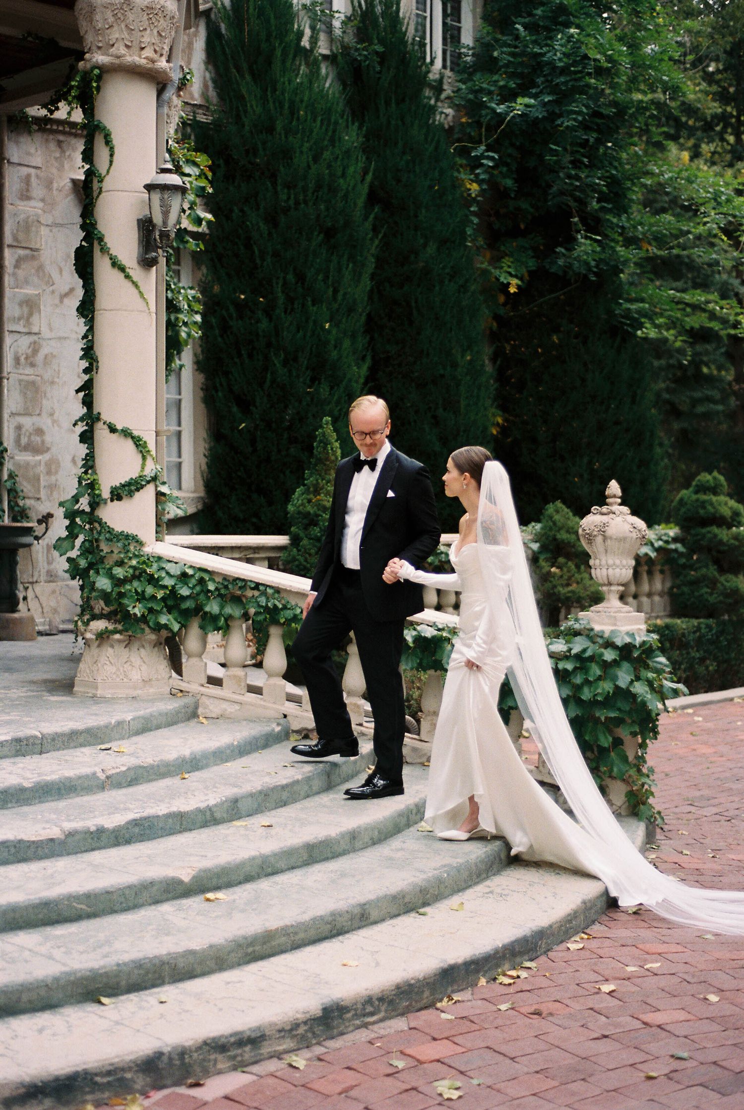 Wedding couple descending ornate stone steps with ivy-covered mansion backdrop.