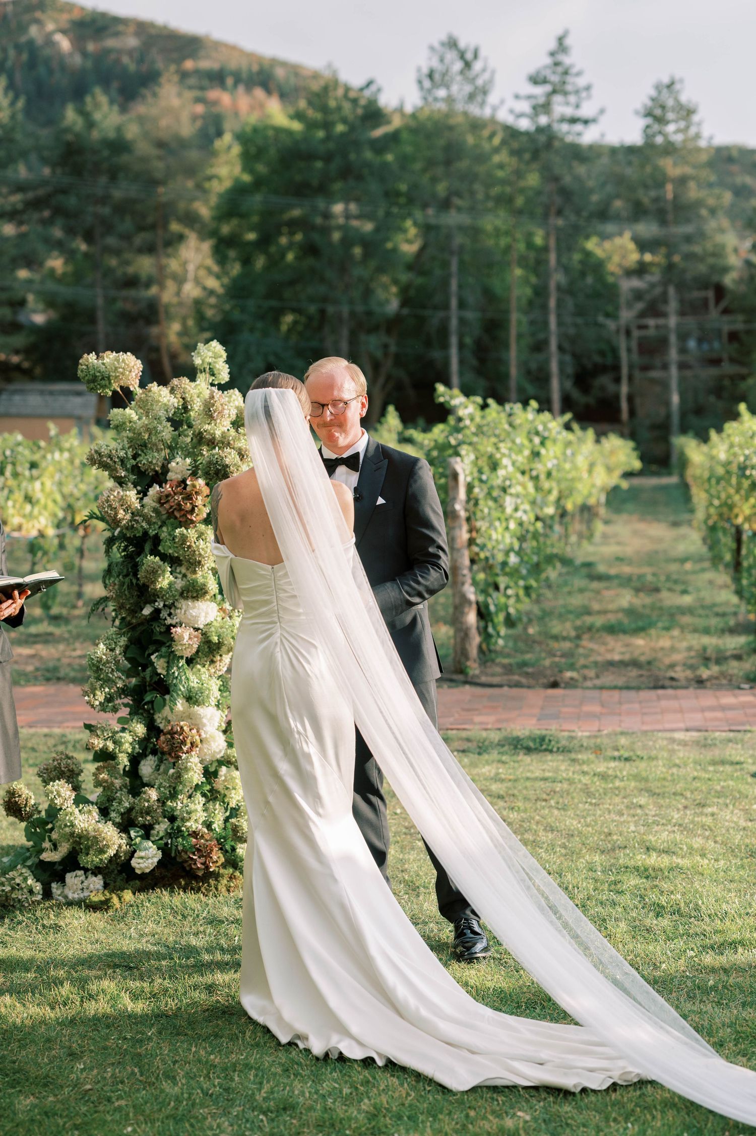 Elegant outdoor wedding ceremony in a vineyard with white floral decorations and dramatic mountain backdrop.