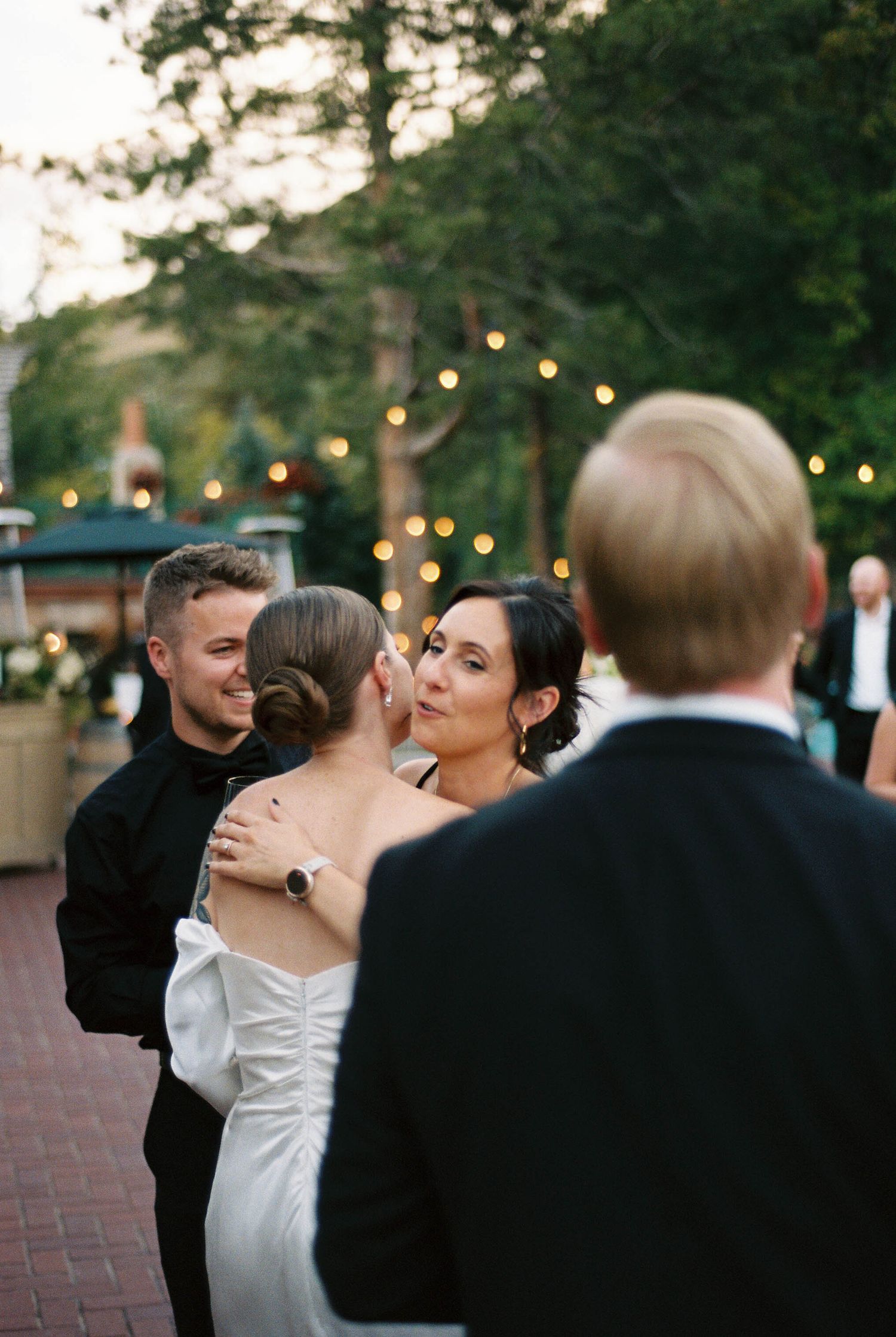 Wedding celebration under string lights with guests in formal attire gathering on brick pavement.