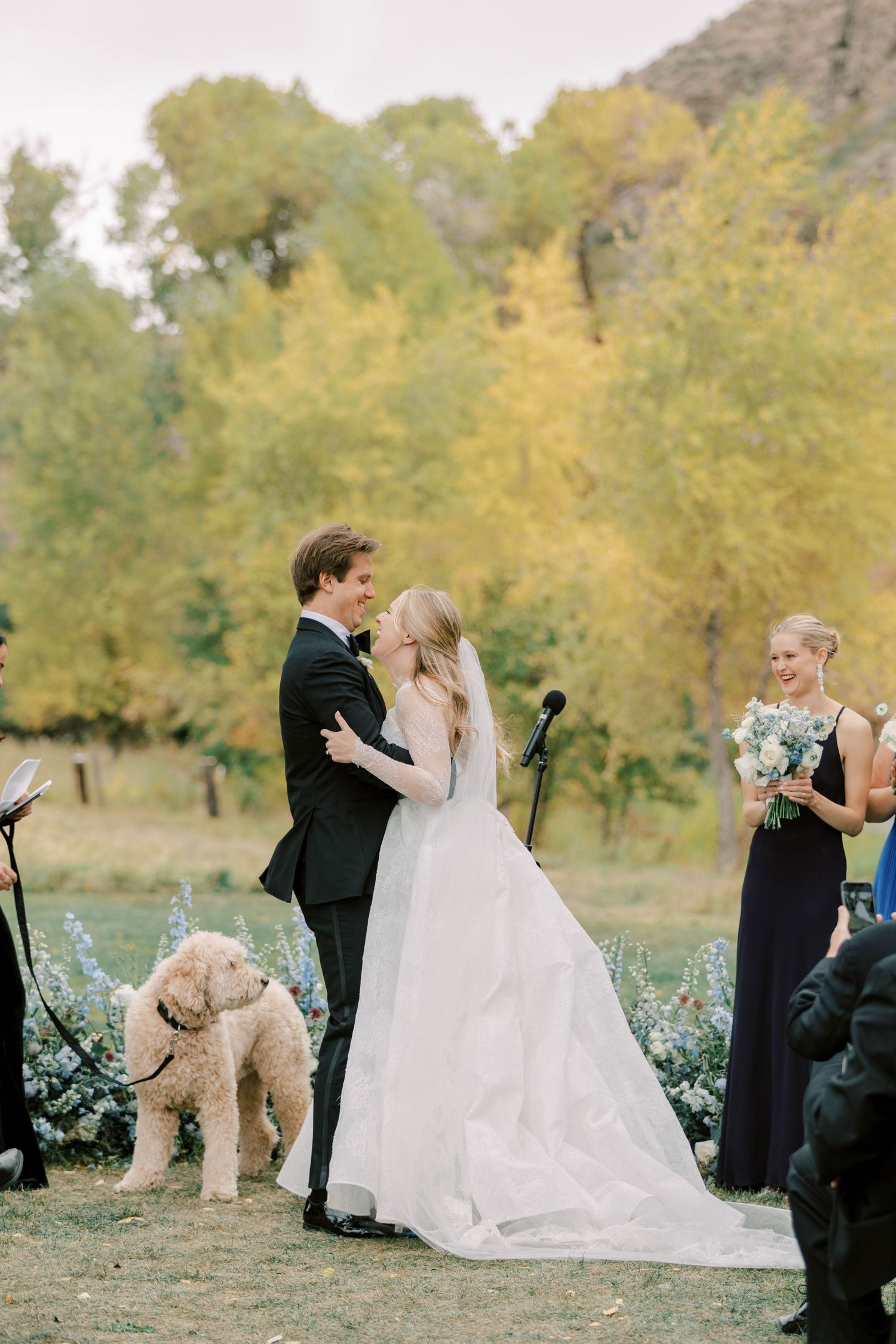 Newlyweds share first kiss at altar with their golden doodle dog and bridesmaid in navy dress nearby.