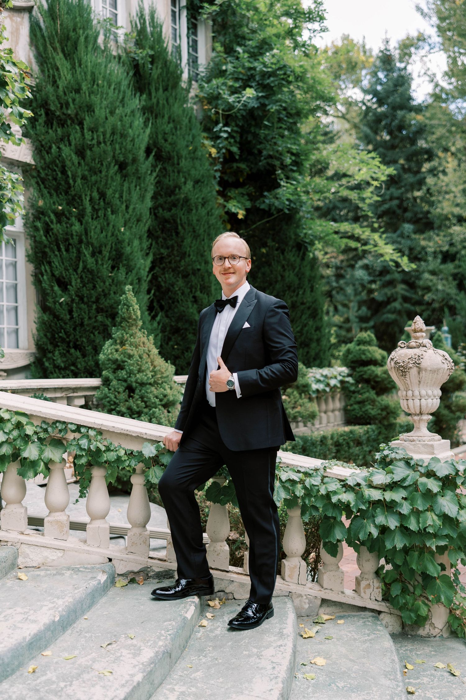 Groom in classic black tuxedo and bow tie poses on elegant stone steps surrounded by greenery.