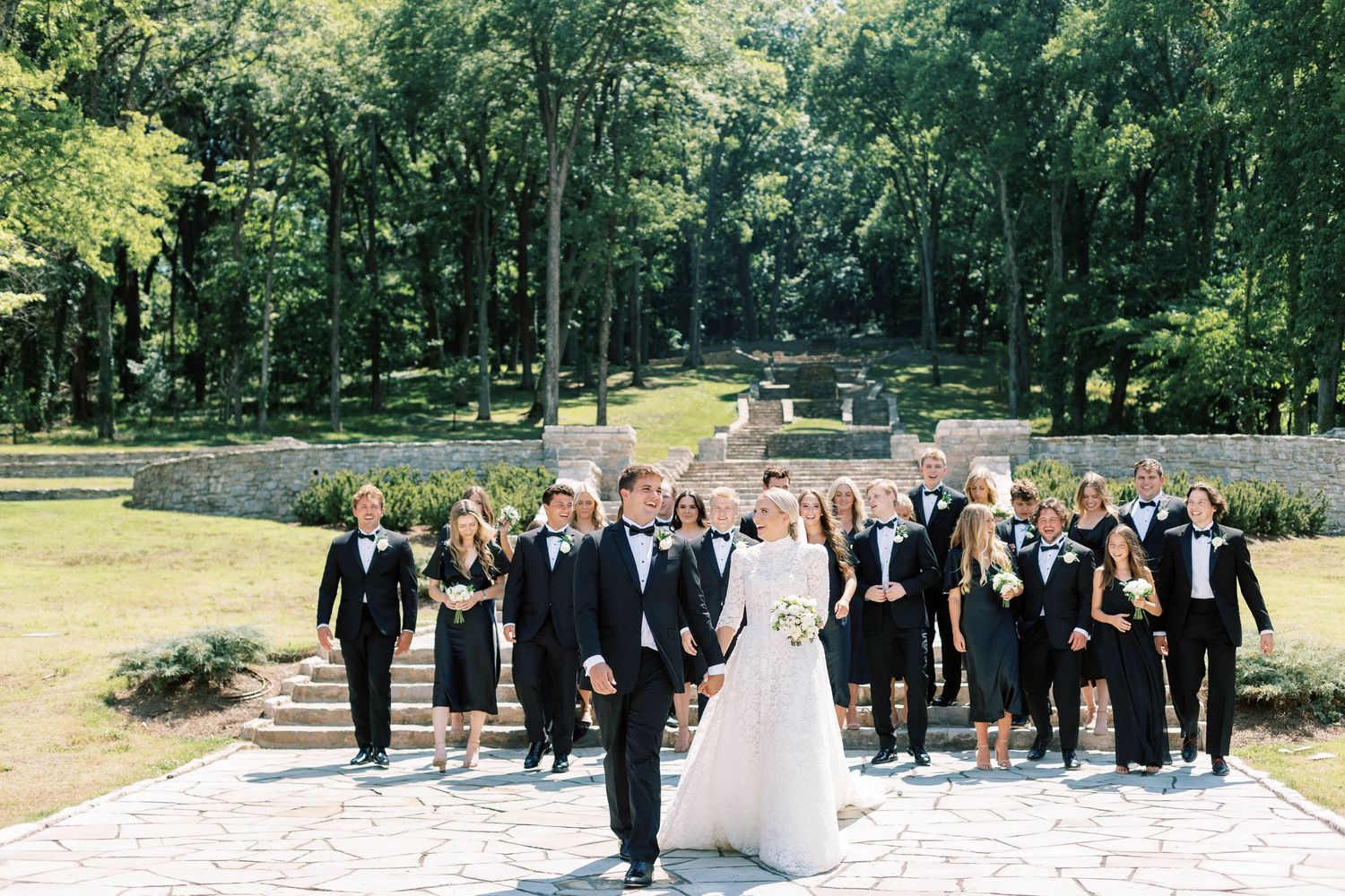 Wedding party walks together on stone path surrounded by lush green trees.