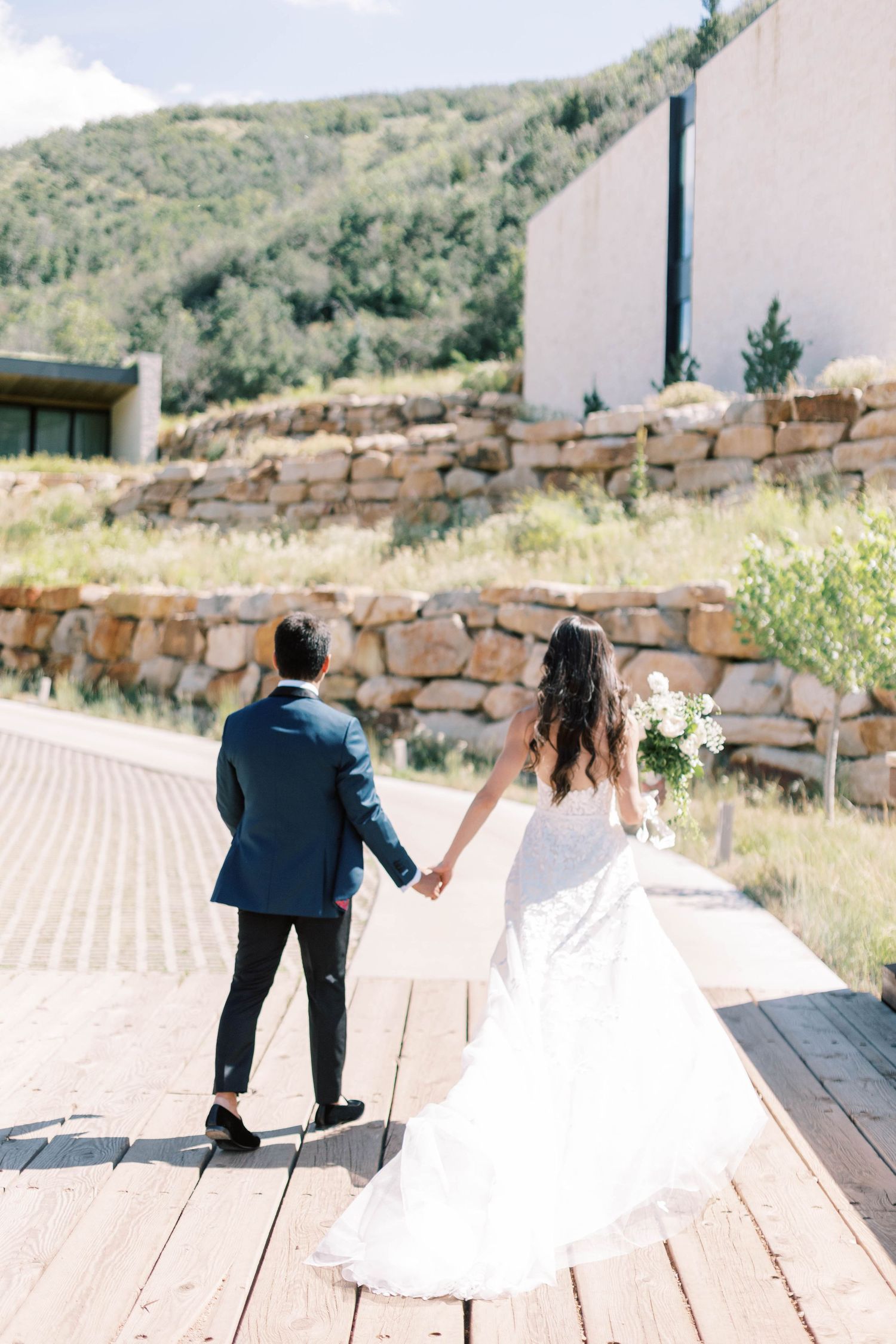modern Newlyweds walking hand in hand on wooden deck with mountain views at blue sky ranch
