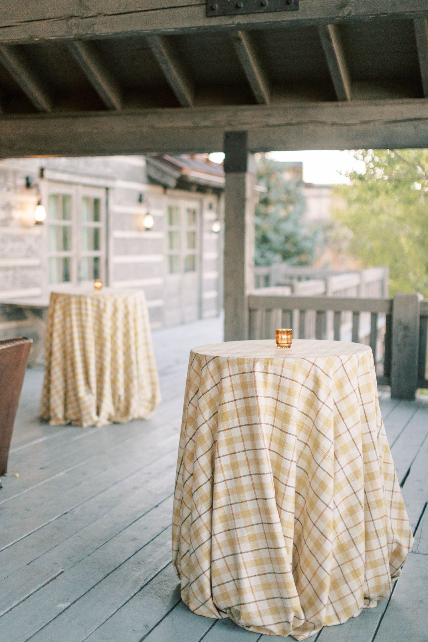 Plaid tablecloths cover cocktail tables on a wooden deck at an outdoor event.