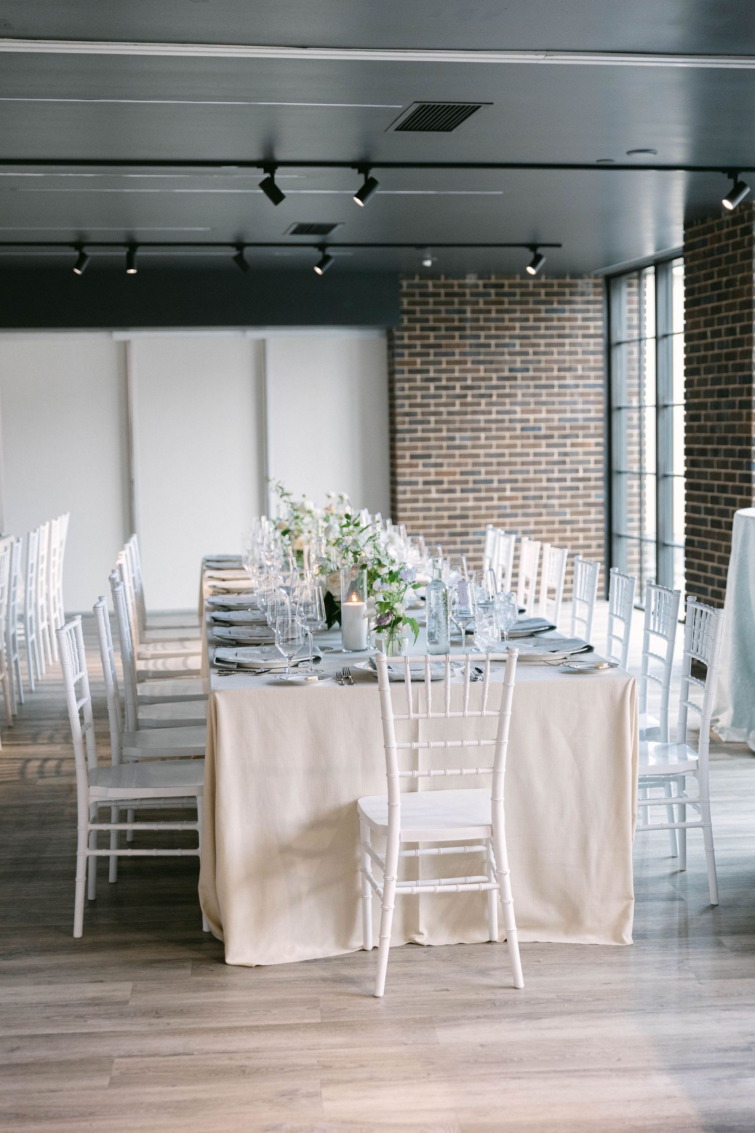 Modern reception venue with white chiavari chairs and cream tablecloths against exposed brick walls.