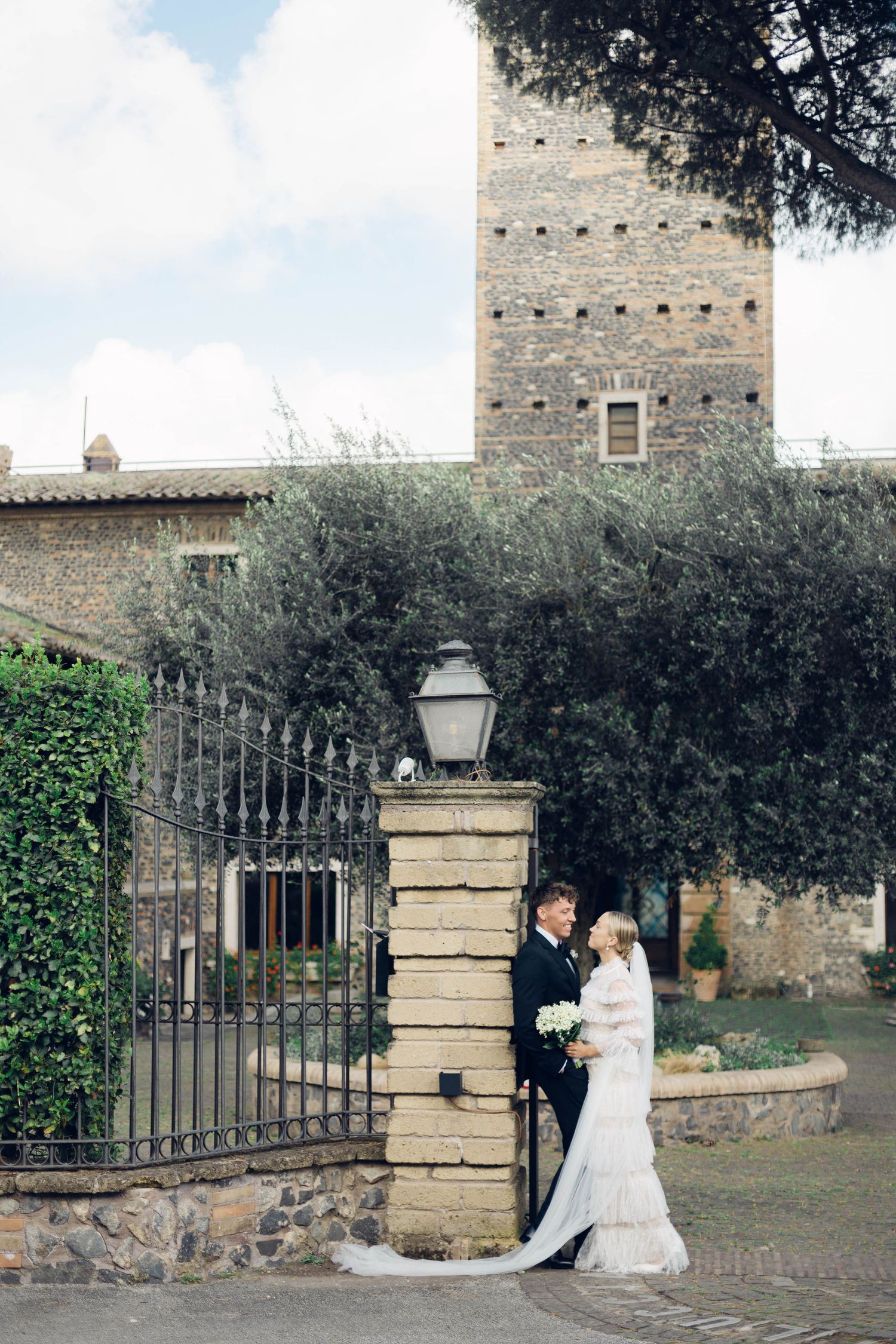 Wedding couple poses by an ancient stone tower and iron gate in a rustic Italian setting at castello della castellucia in rome.