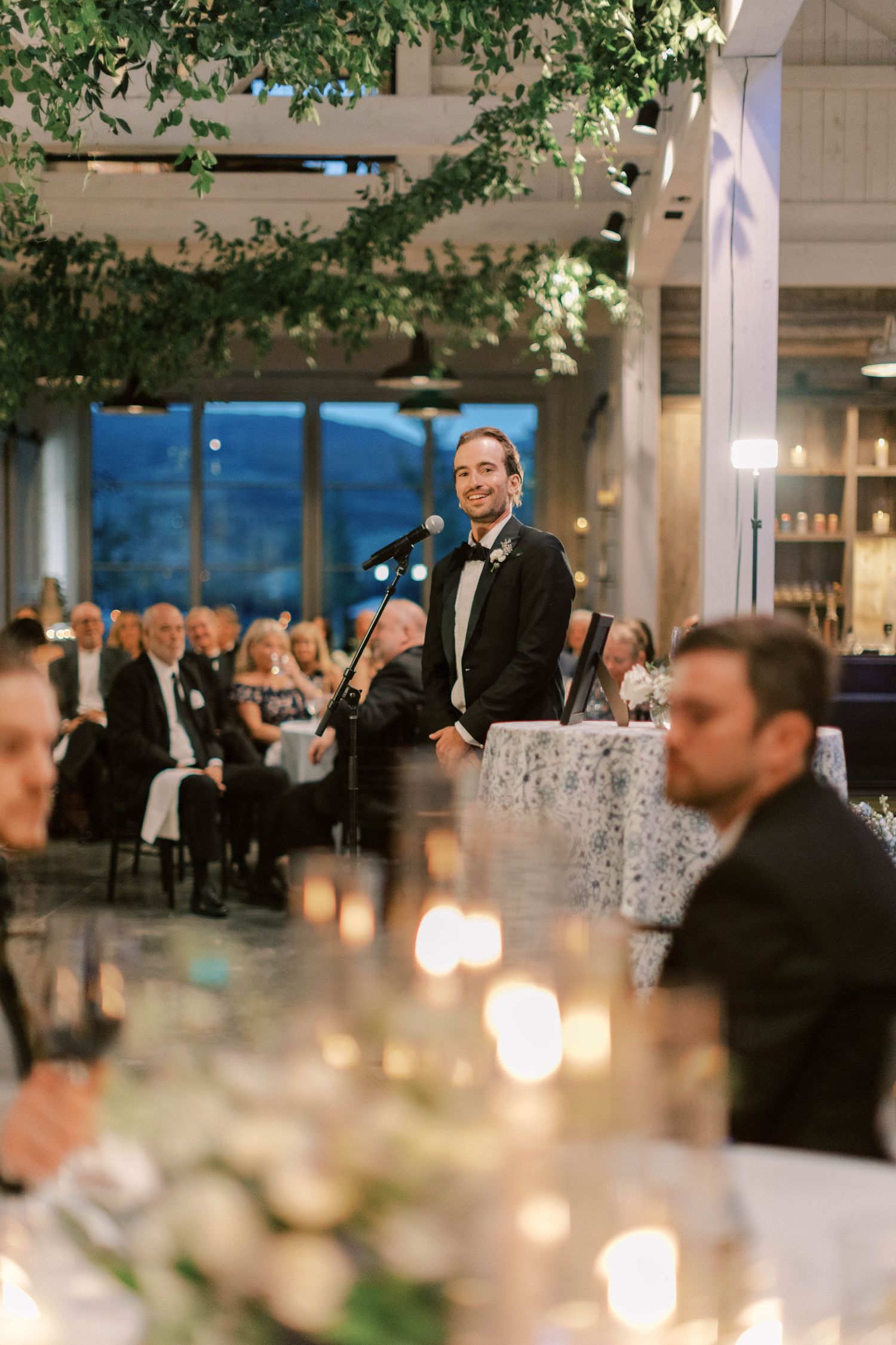 Someone delivers a wedding speech at an elegant indoor reception with candlelit tables and greenery decorations.