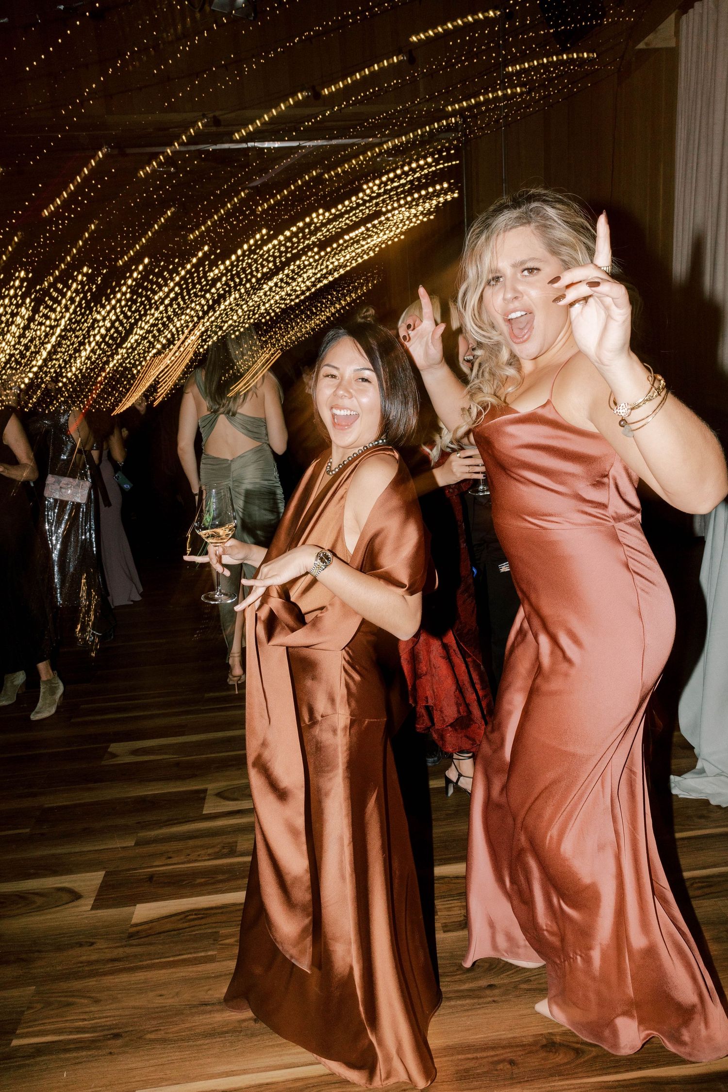 Party guests in silk dresses dancing and celebrating under twinkly string lights at an elegant evening reception.