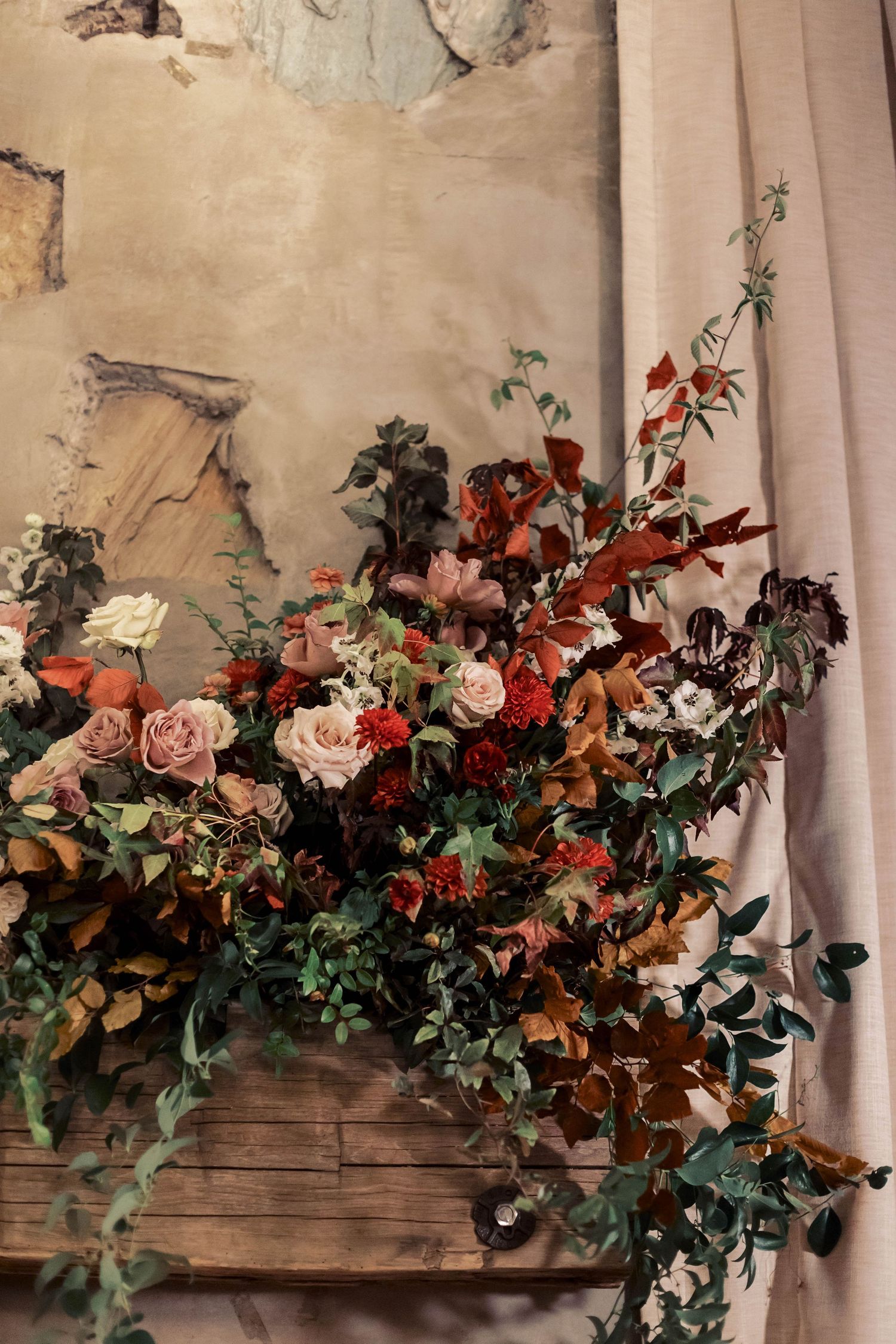 Lush floral arrangement with red and pink roses, autumn leaves, and greenery against a rustic wall and curtain.