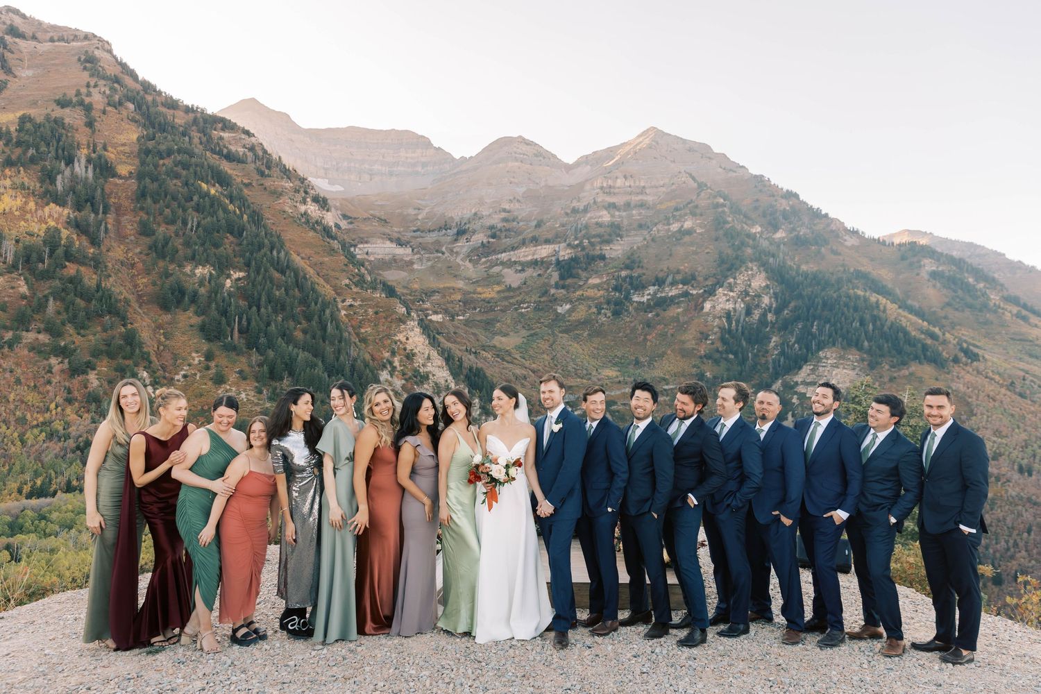 Wedding party in navy suits and jewel-toned dresses pose in front of mountain range.
