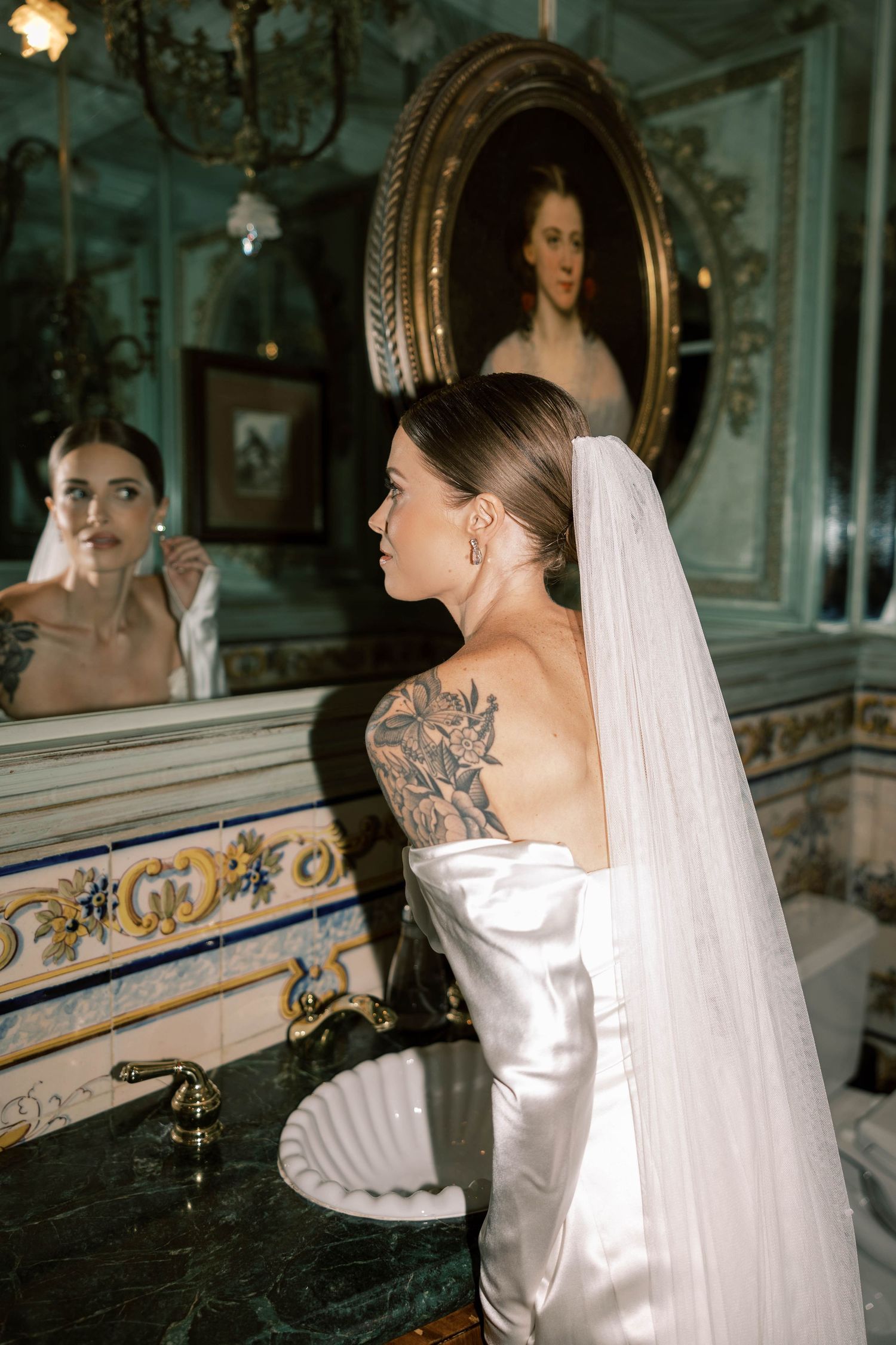 Bride in silk dress with shoulder tattoo looks in vintage mirror of elegant room.