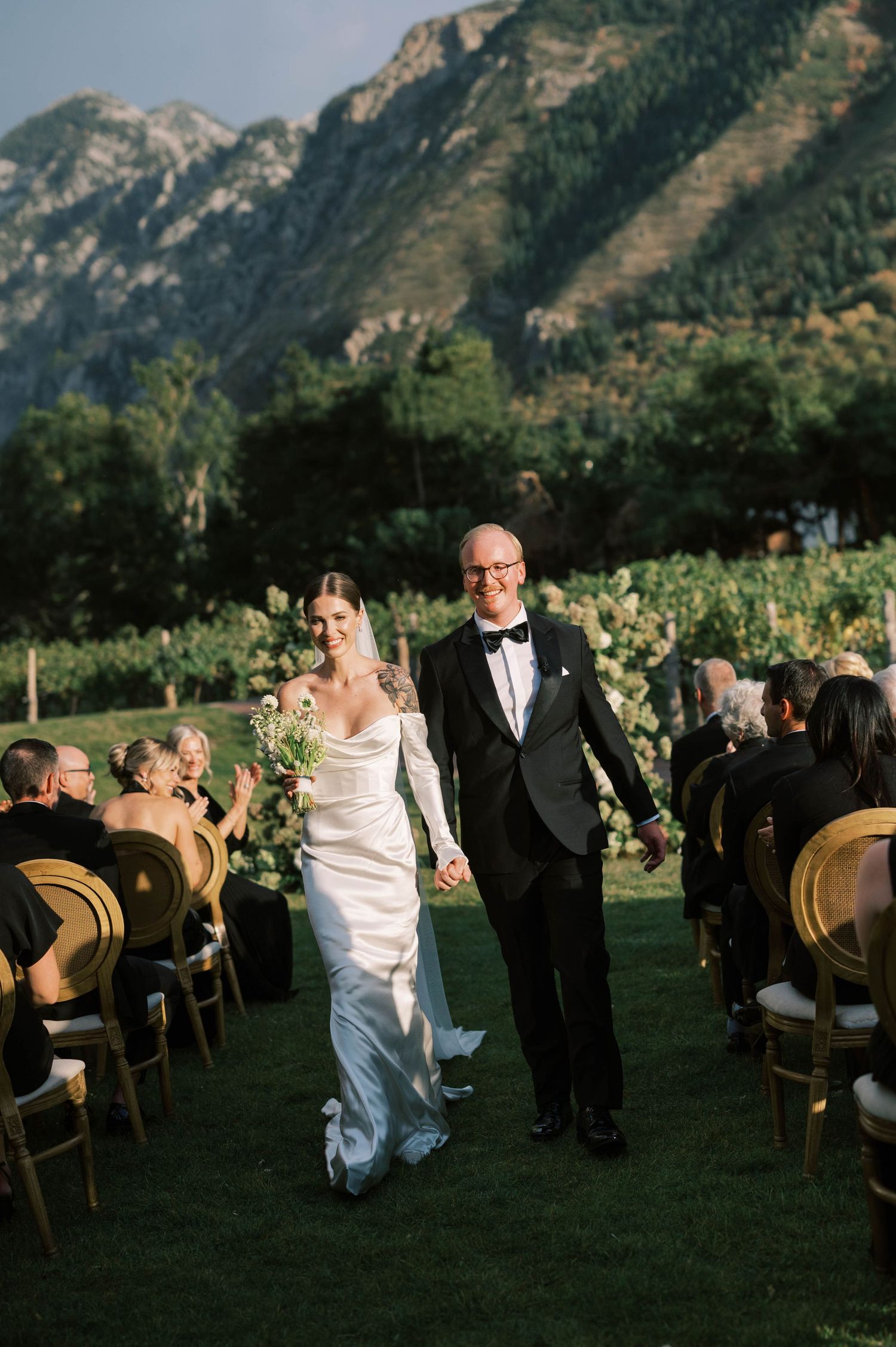 A wedding ceremony takes place outdoors with majestic mountains in the background as guests watch from wooden chairs.
