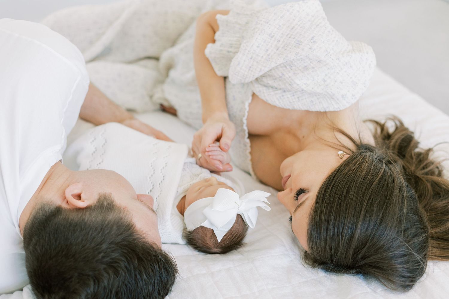 Family lying together on white bedding while holding newborn baby.