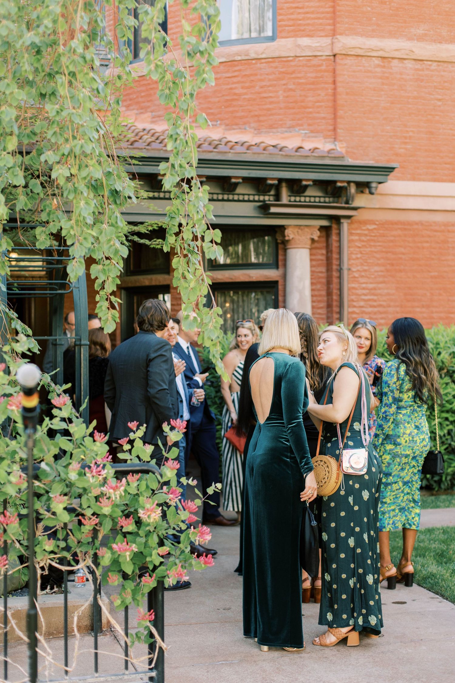 Wedding guests in formal attire gather outside brick building among garden foliage.