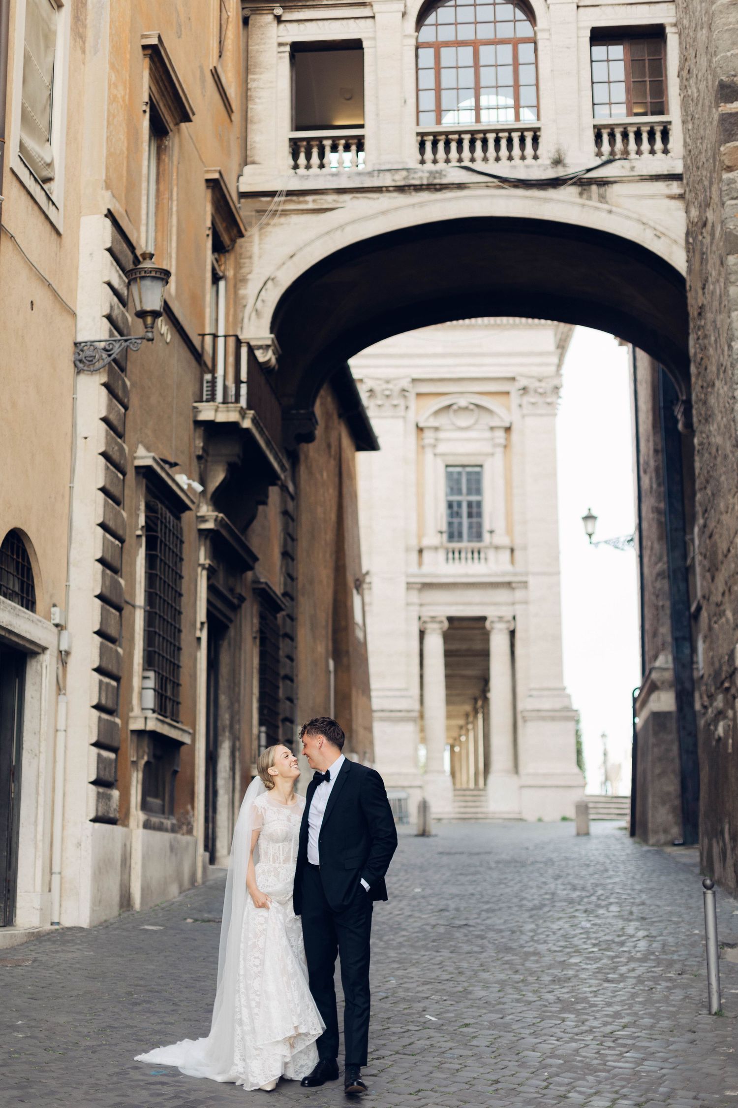 Wedding couple stands beneath ornate stone archway on cobblestone street in Rome.