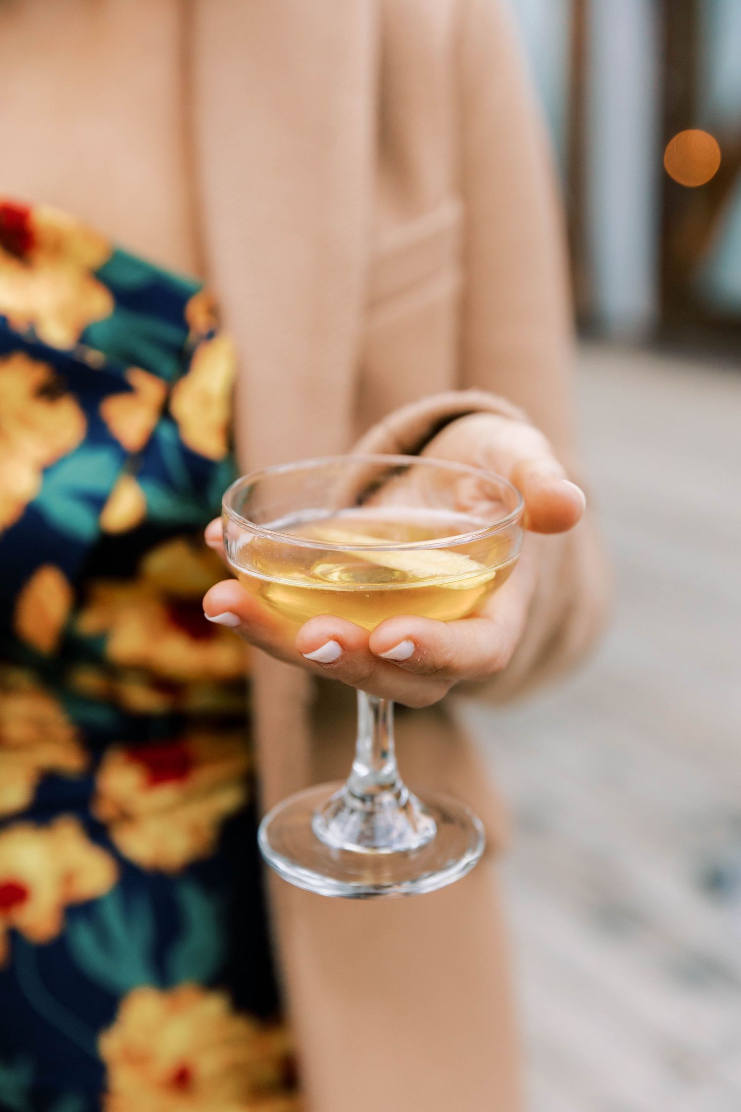 Hand holding a coupe glass filled with golden champagne against a floral dress background.