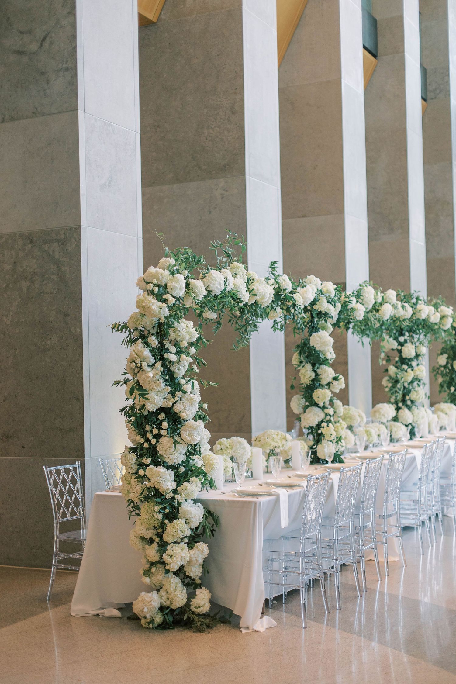 Elegant wedding reception table with lush white floral arch and ghost chairs in a modern columned venue.