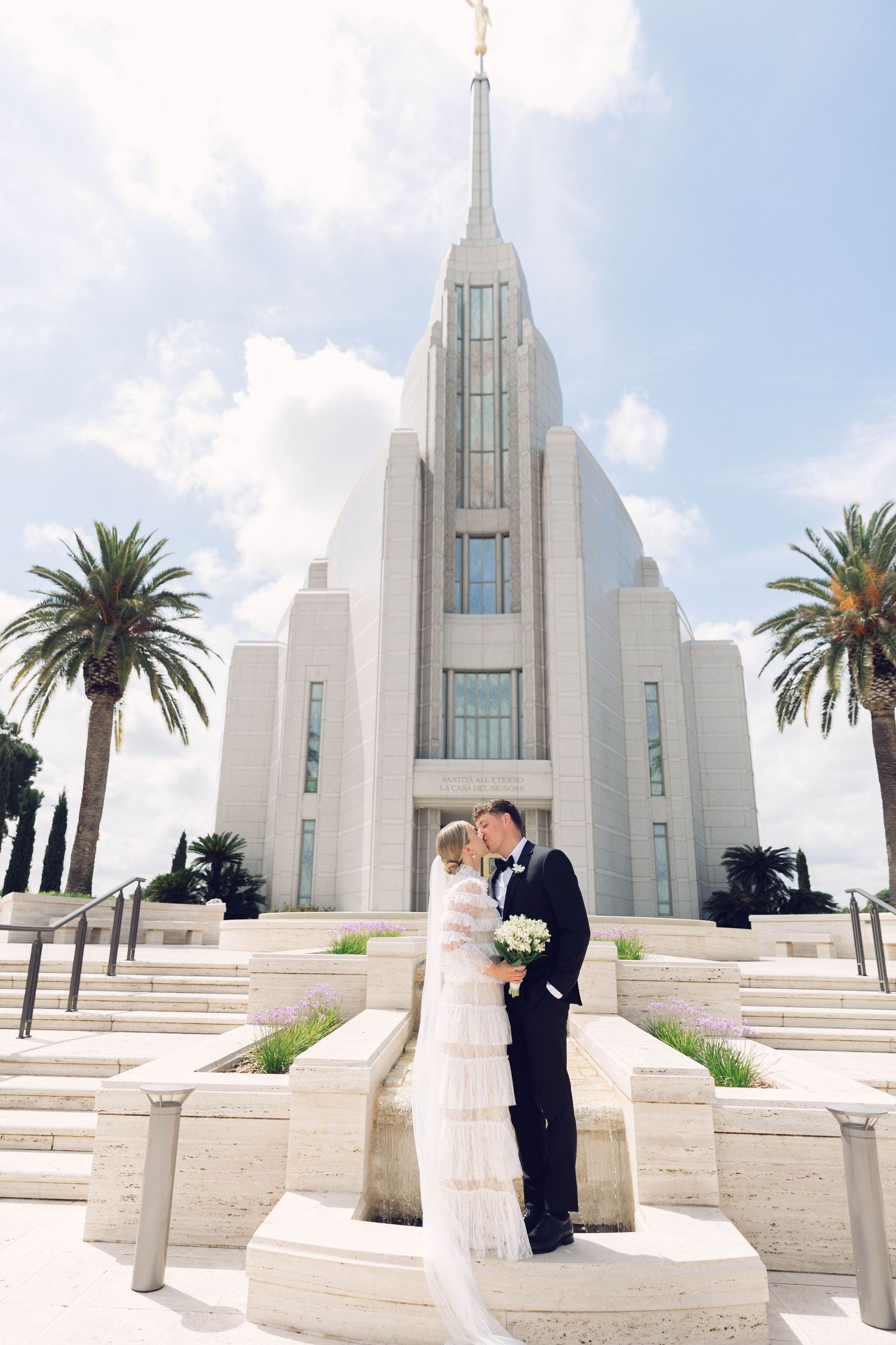 Wedding couple posing on temple steps with white architecture and palm trees in background at rome italy lds temple.
