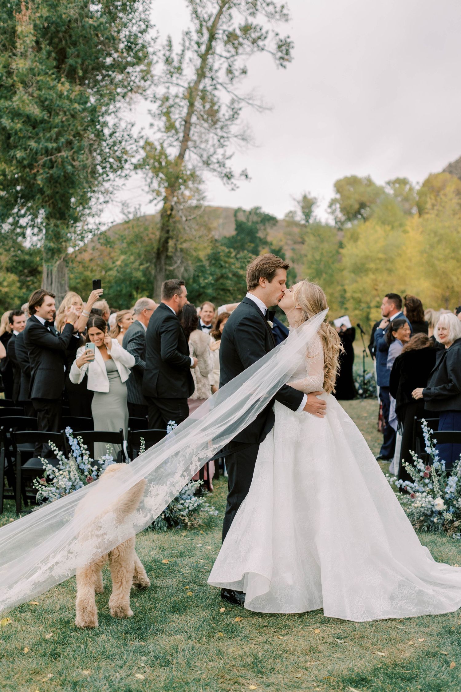 Bride and groom share first kiss as newlyweds while their golden doodle watches during outdoor ceremony.