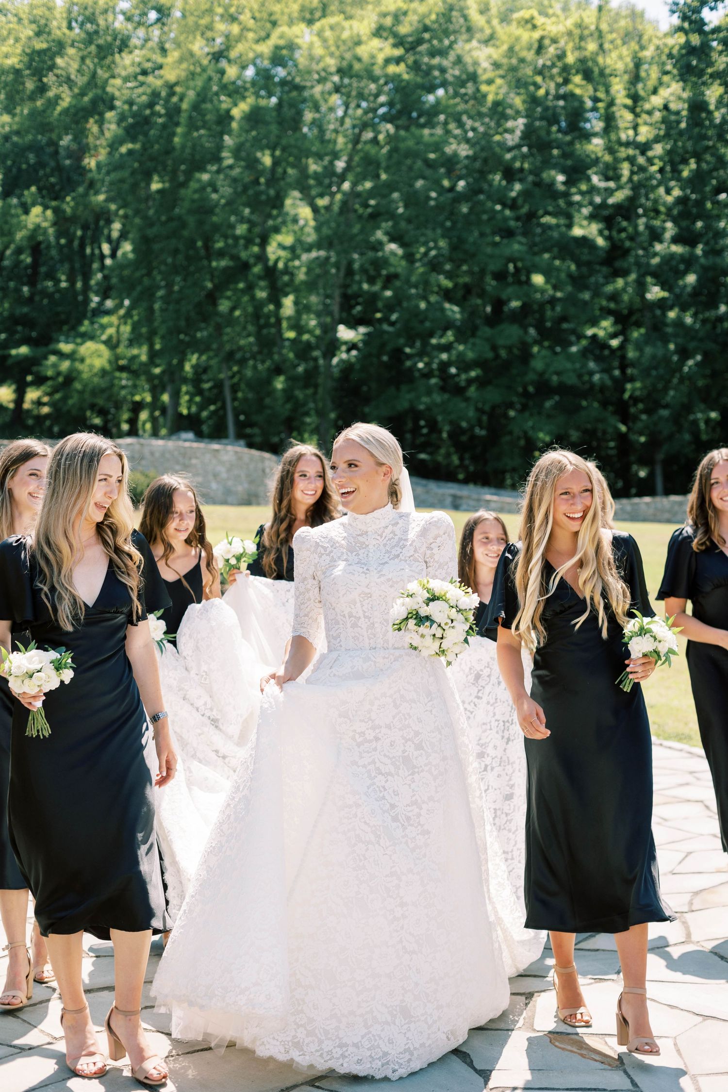 Bride in lace gown stands with bridesmaids wearing black dresses in outdoor garden setting.