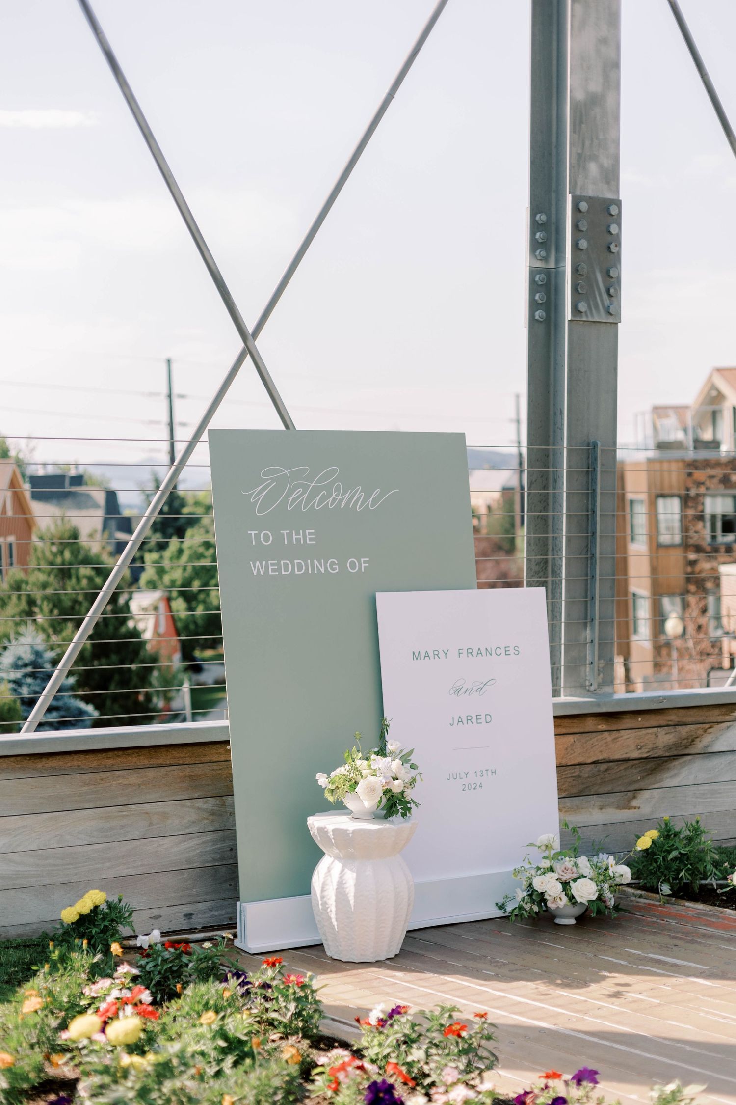 Wedding welcome sign display with sage green boards and white floral arrangements on an outdoor deck.