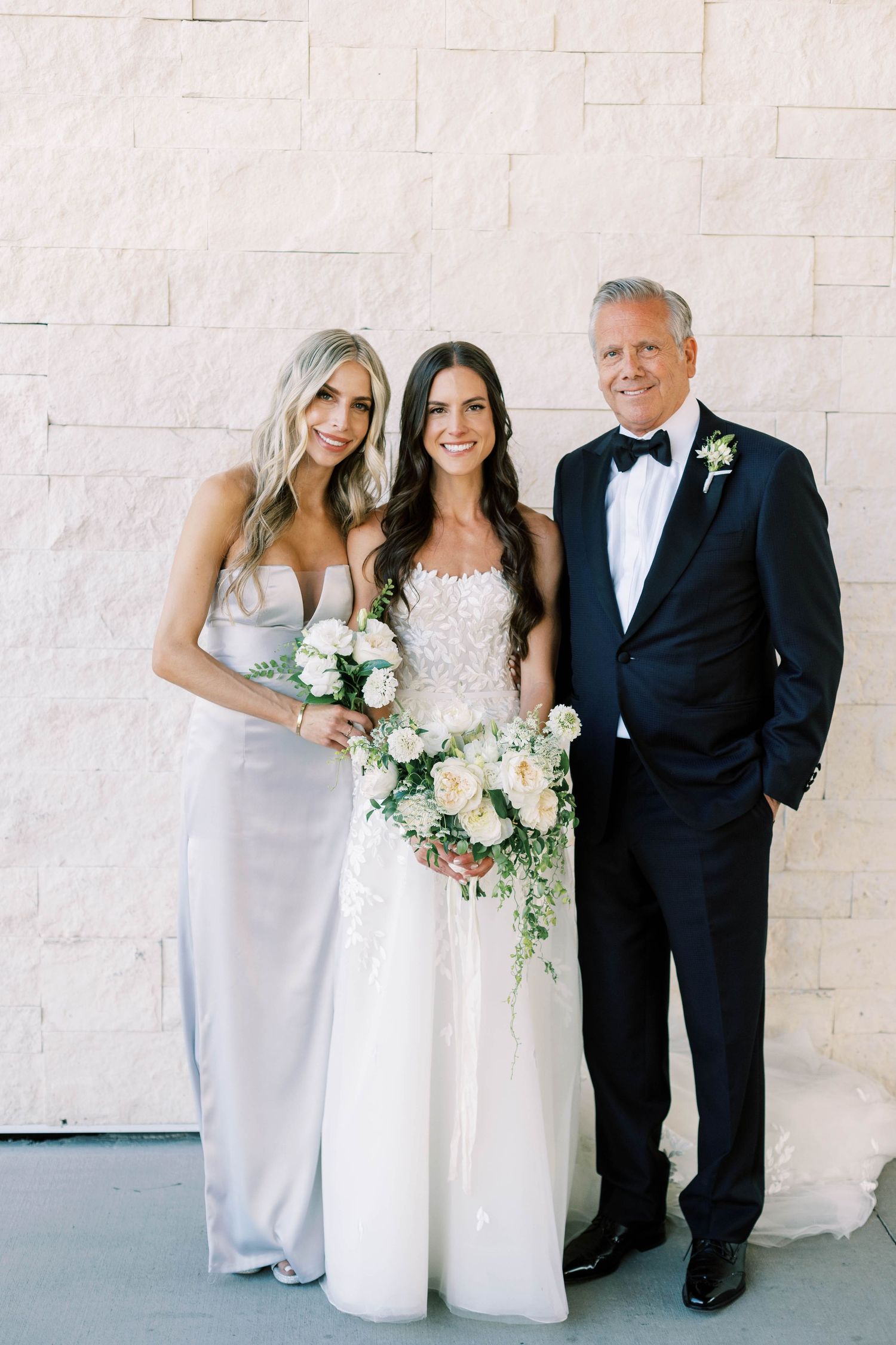 Family formal portrait with bride and wedding party members against white stone wall.