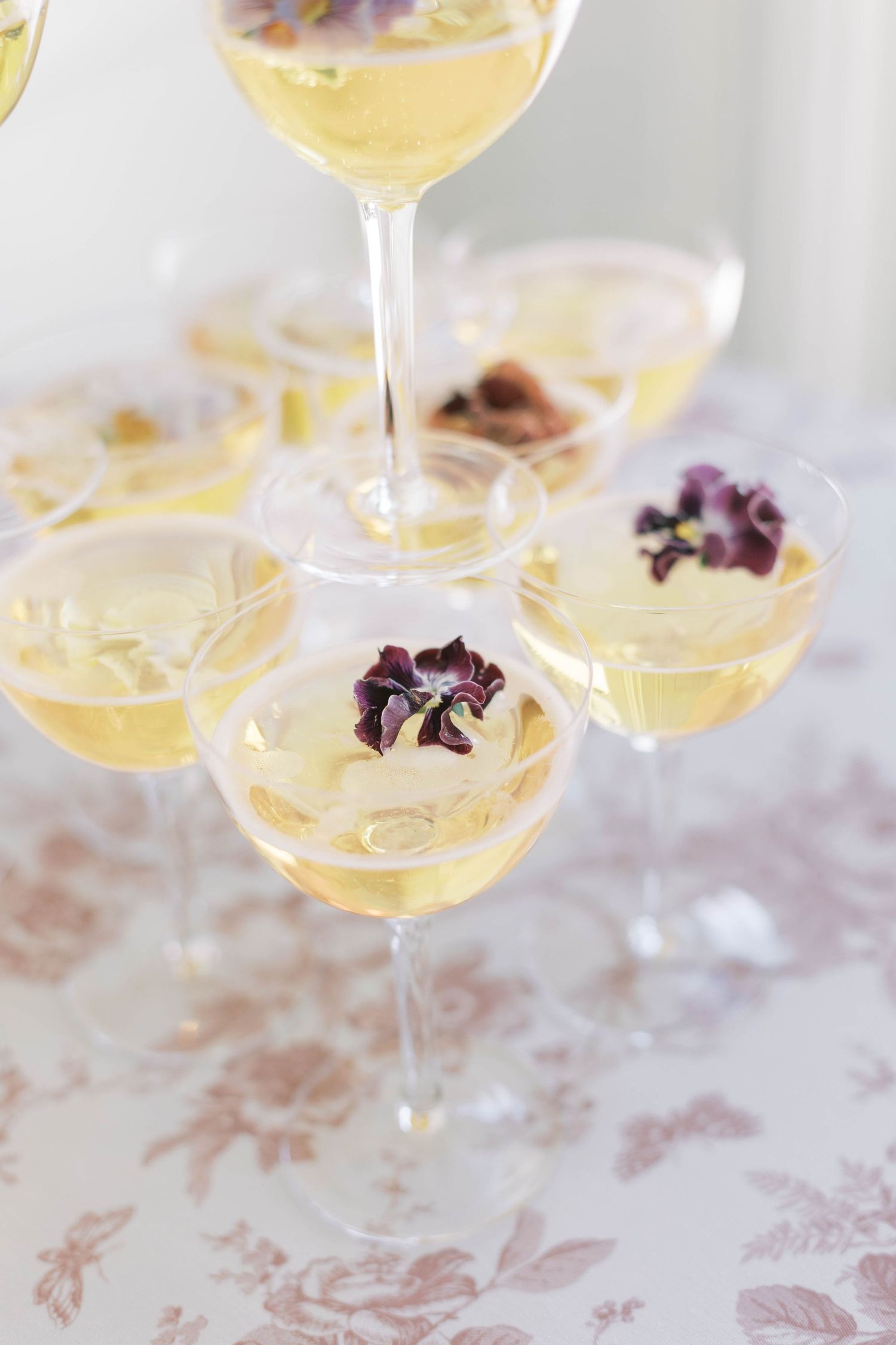 Champagne coupe glasses filled with yellow cocktails garnished with purple edible flowers.