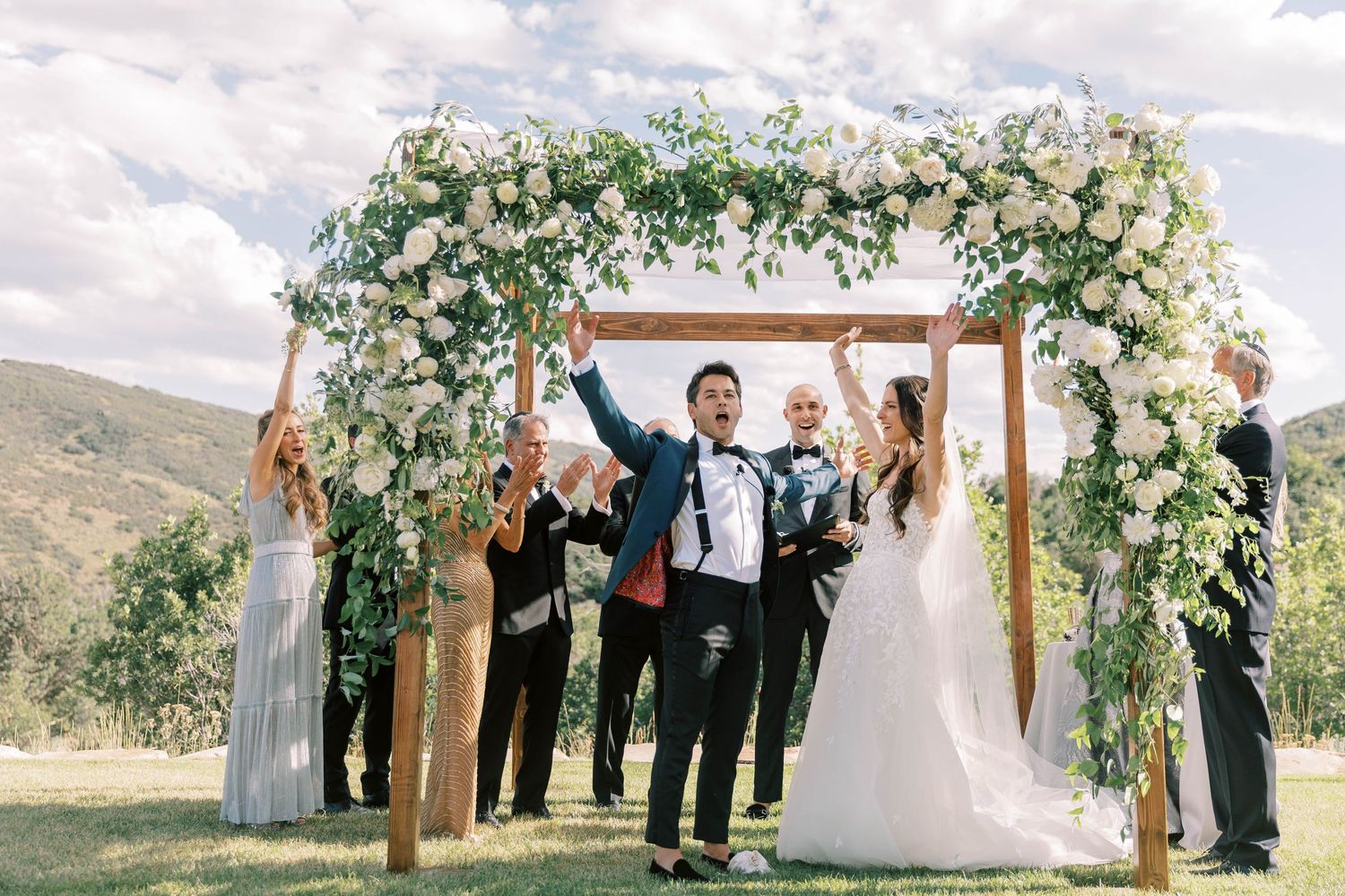 Wedding ceremony under a floral arch with mountains in the background on a sunny day.