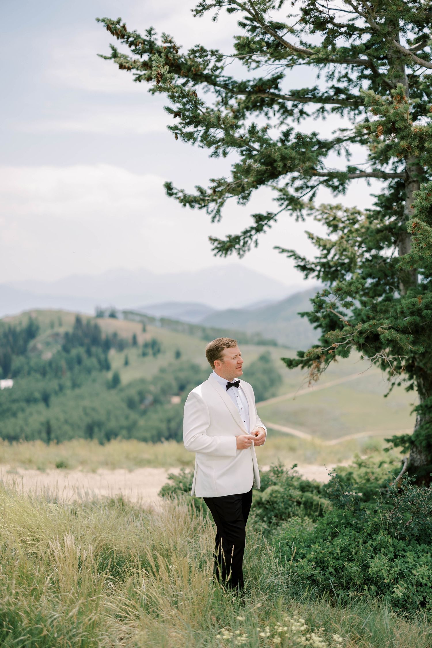 Groom in white dinner jacket stands on mountainside overlooking scenic valley.
