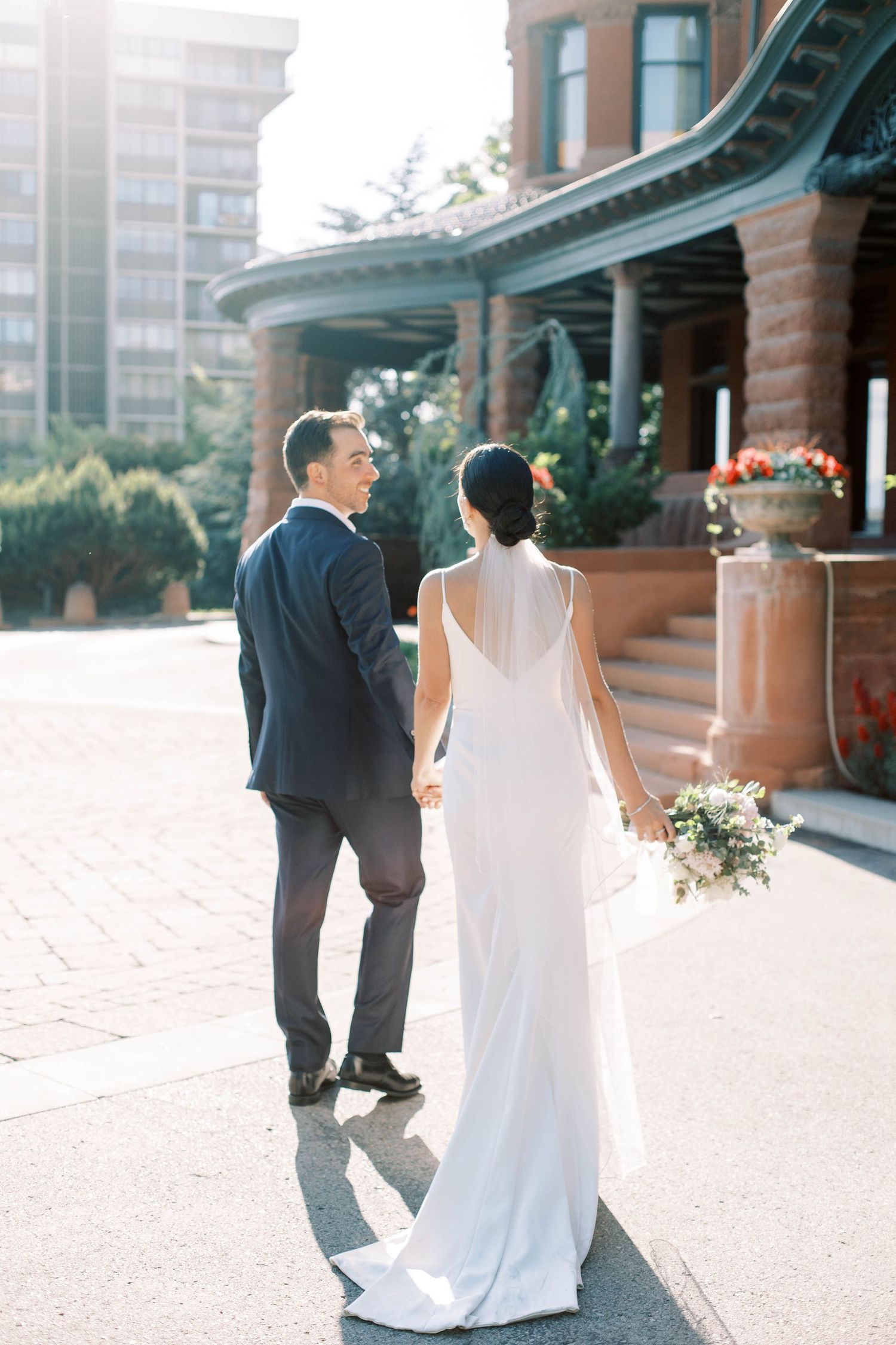 Elegant couple in wedding attire walking together on brick path outside historic building with curved architecture at the mccune mansion.