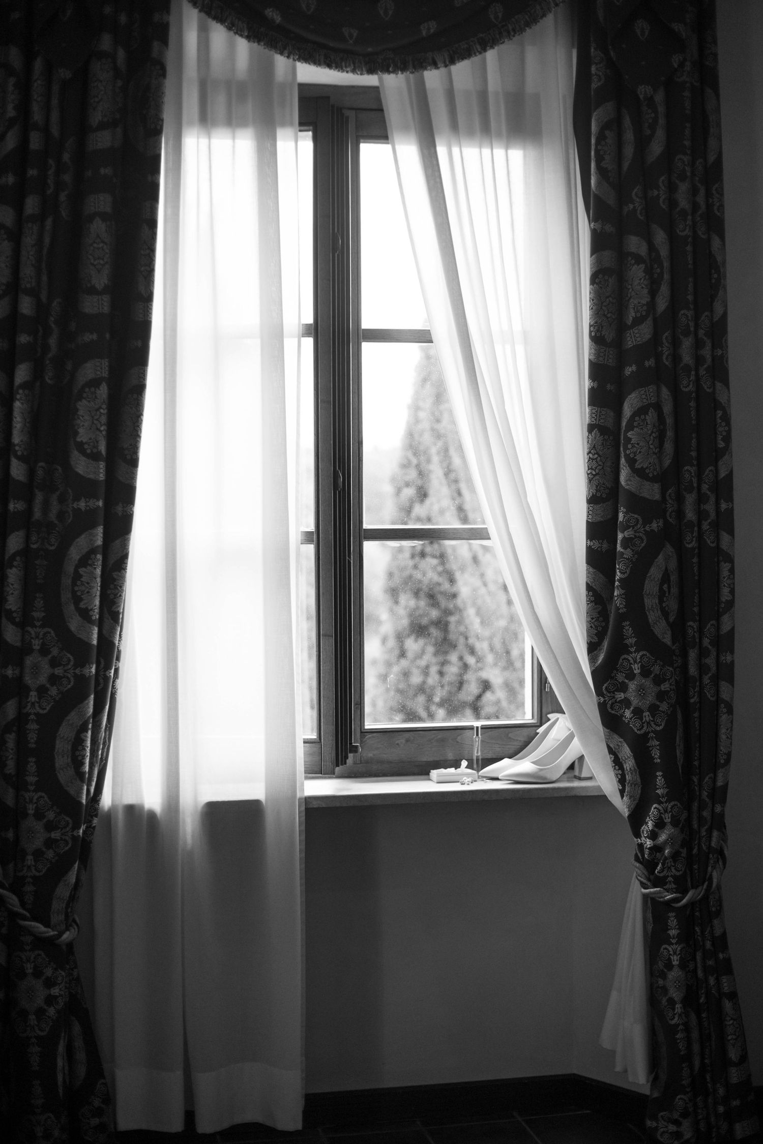 Black and white photograph of window with flowing sheer curtains and cypress trees view.