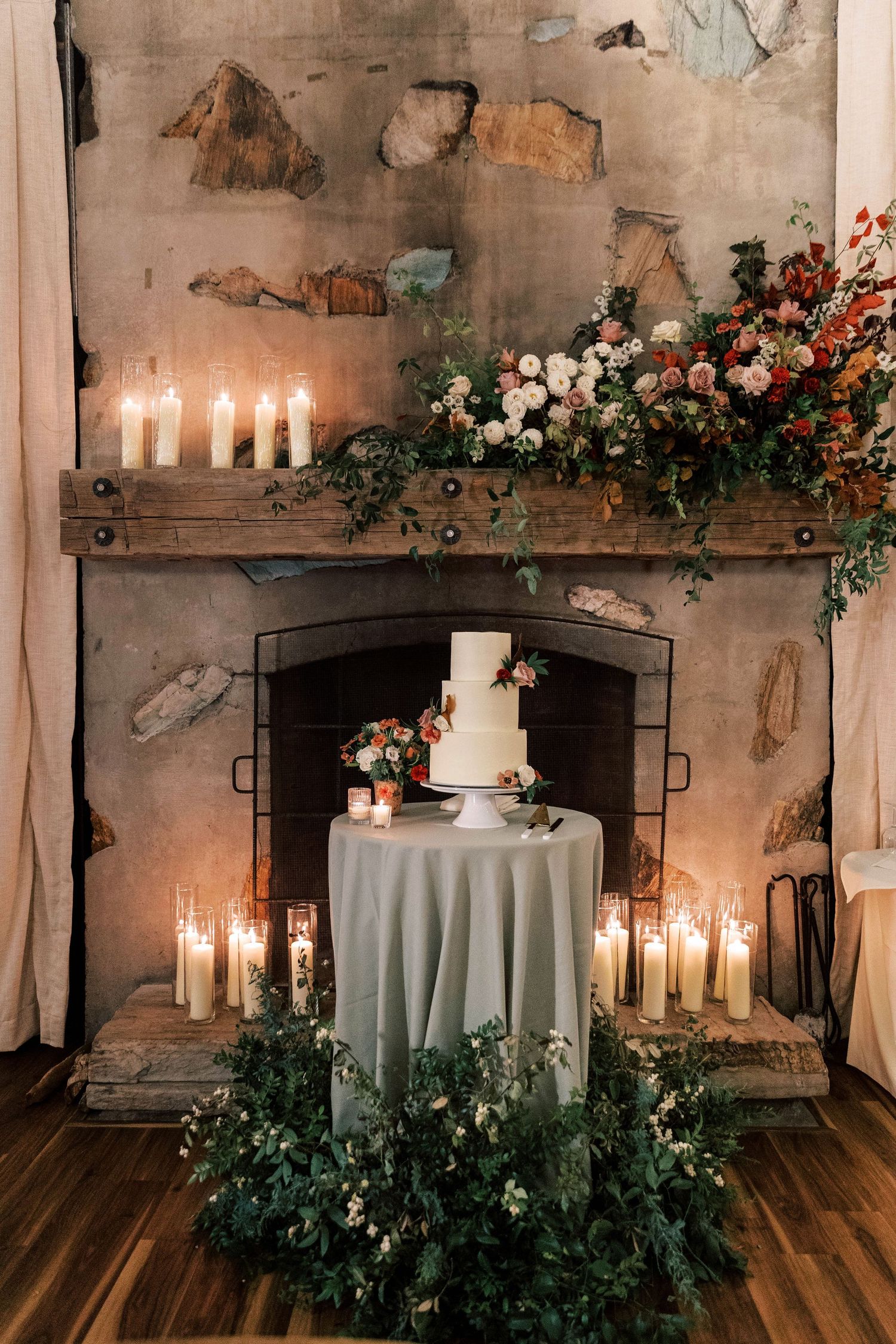 Wedding cake displayed on a round table surrounded by candles and floral arrangements in a rustic fireplace setting.