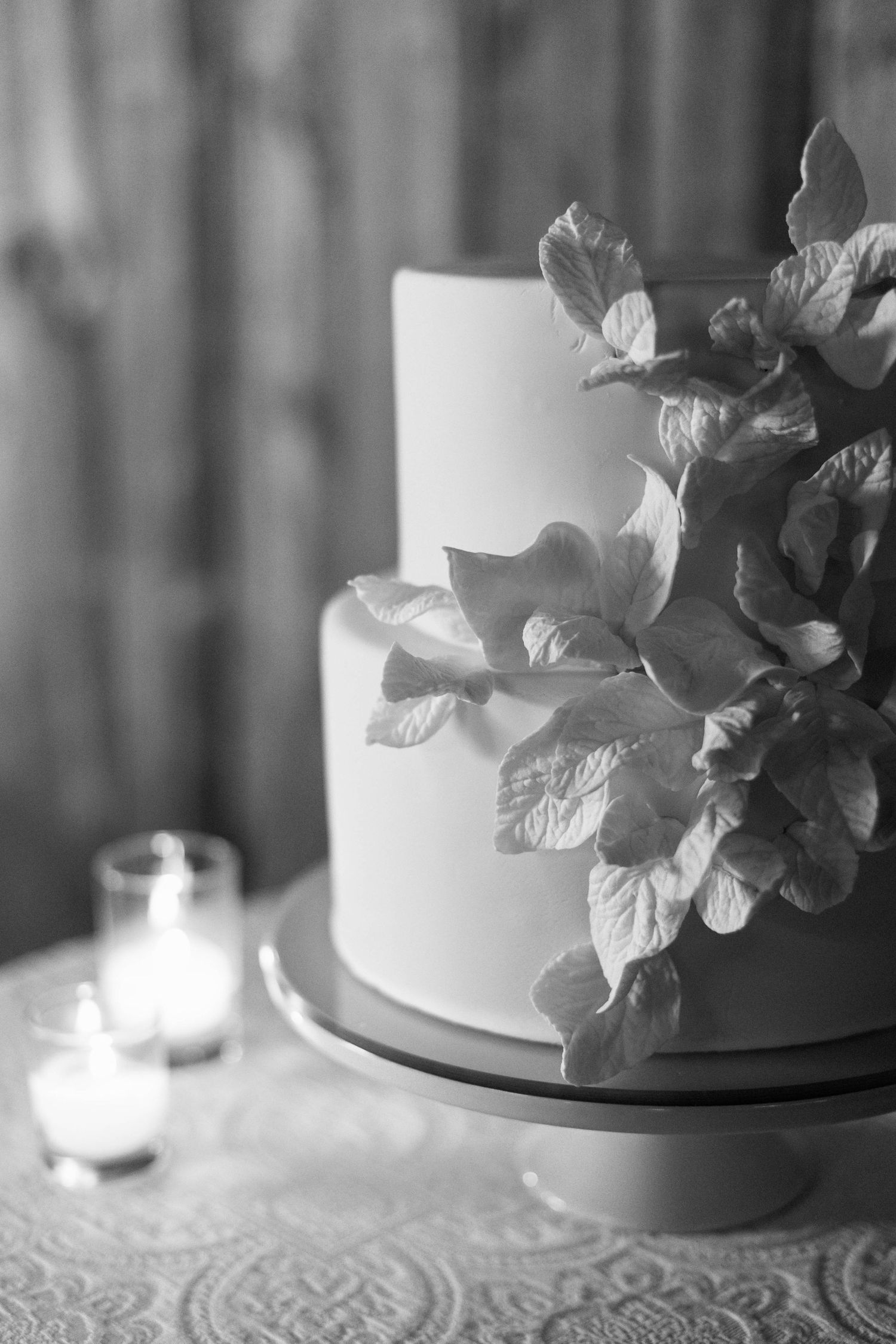 Black and white photo of elegant wedding cake with floral decorations and candlelight.