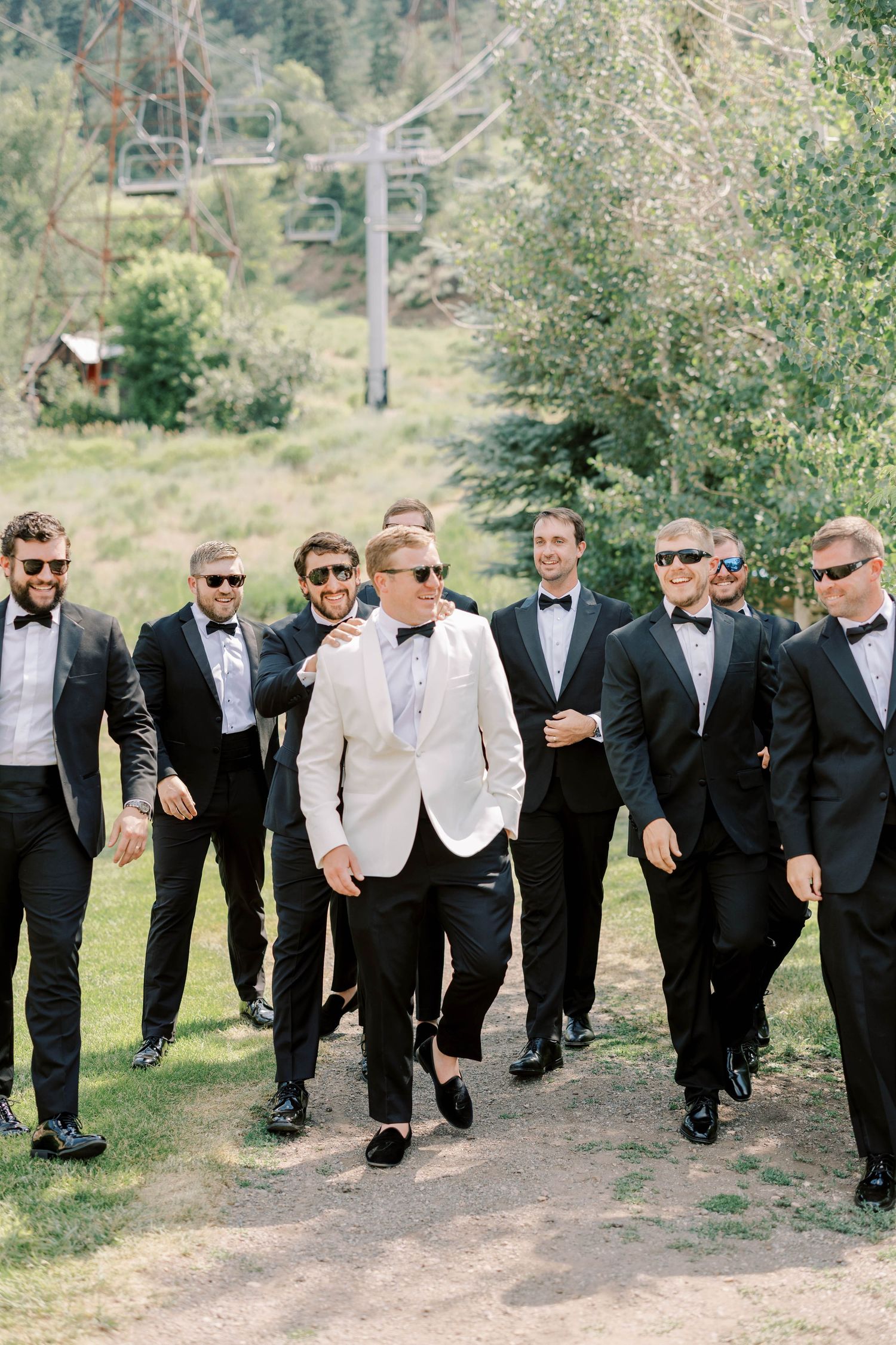 Groomsmen in black tuxedos walk with groom in white jacket on mountain path.