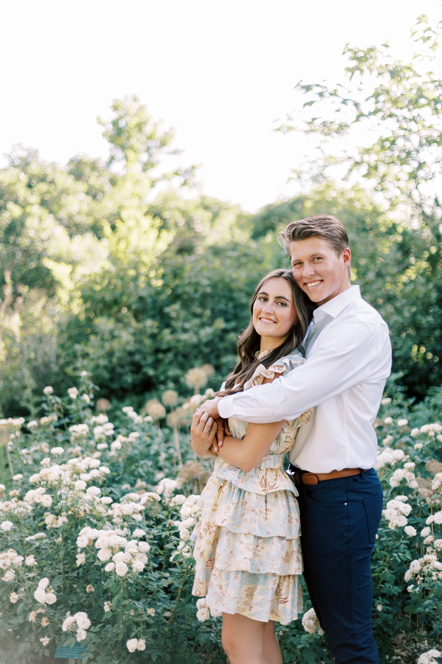 A romantic couple embraces in a garden filled with blooming white flowers and lush greenery.