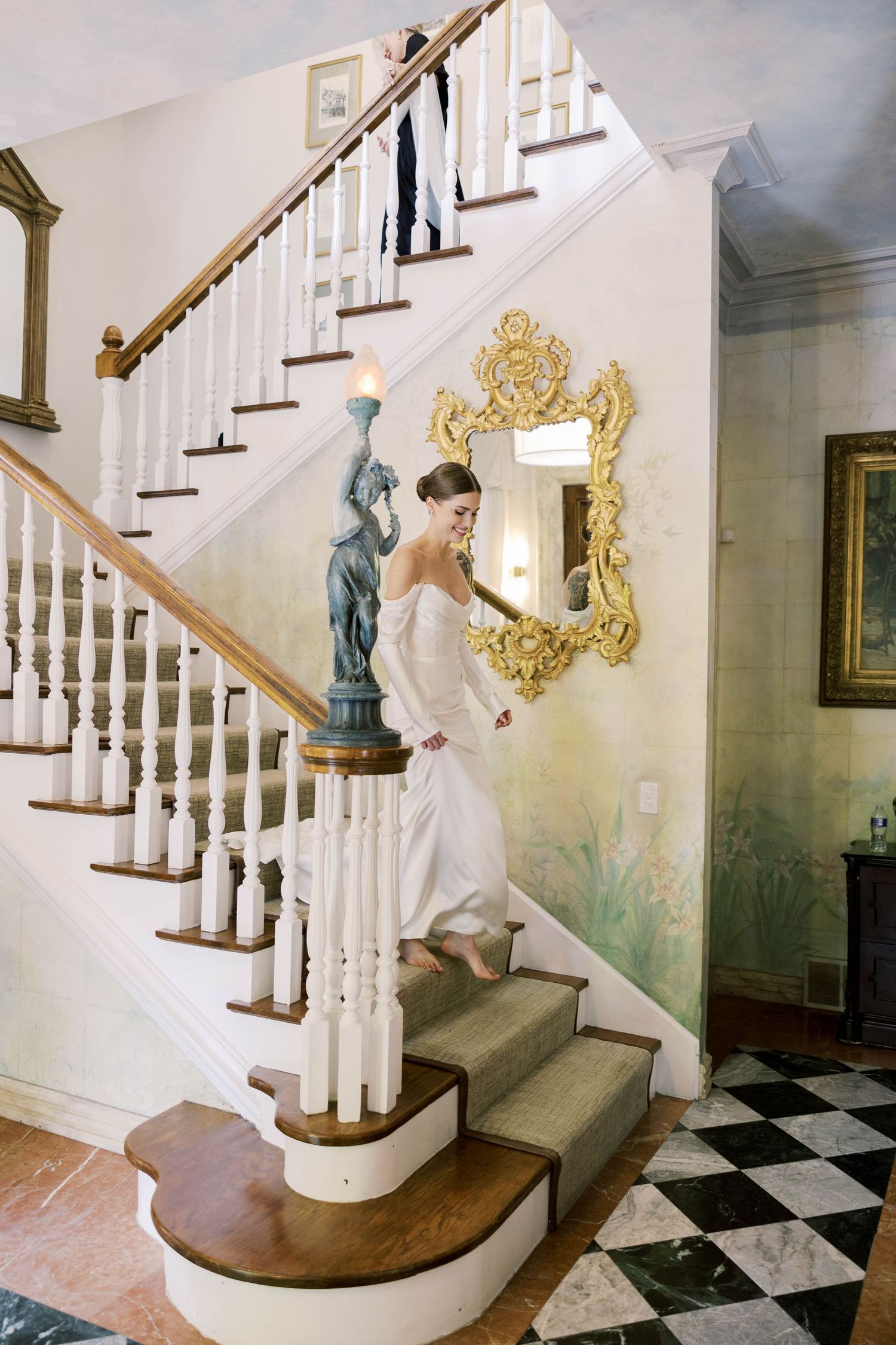 Bride ascending white staircase next to ornate gold mirror in elegant interior.