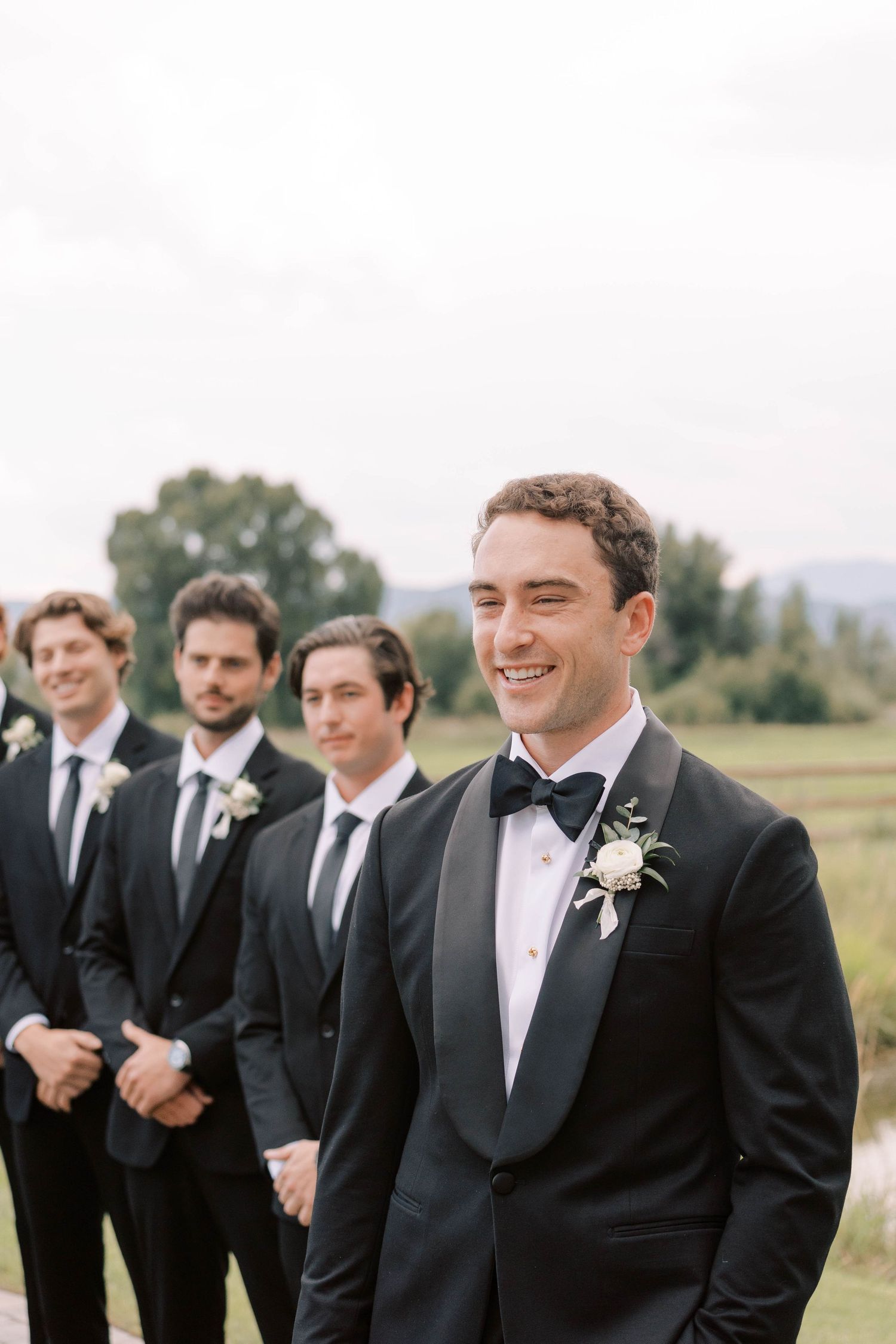 Groomsmen in formal black tuxedos at outdoor wedding venue.