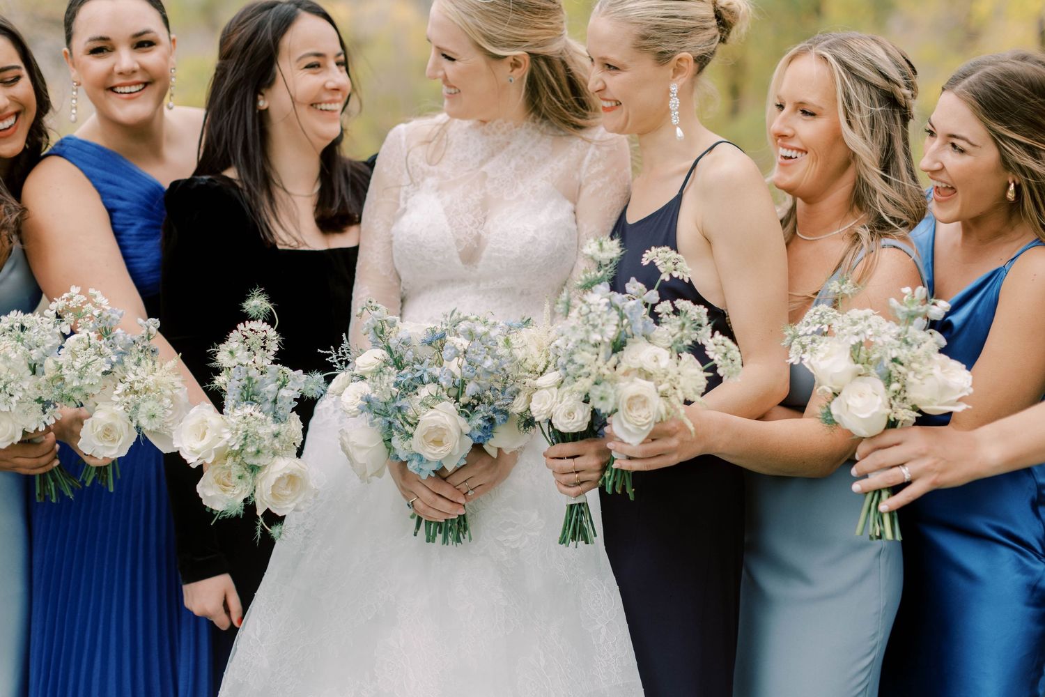Bridal party wearing blue dresses and holding white bouquets laugh together during outdoor wedding photos.