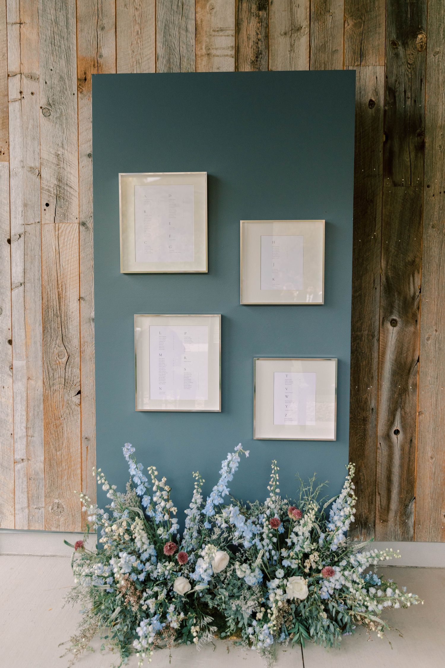 Wedding seating chart display with white frames on teal wall above delicate floral arrangement.