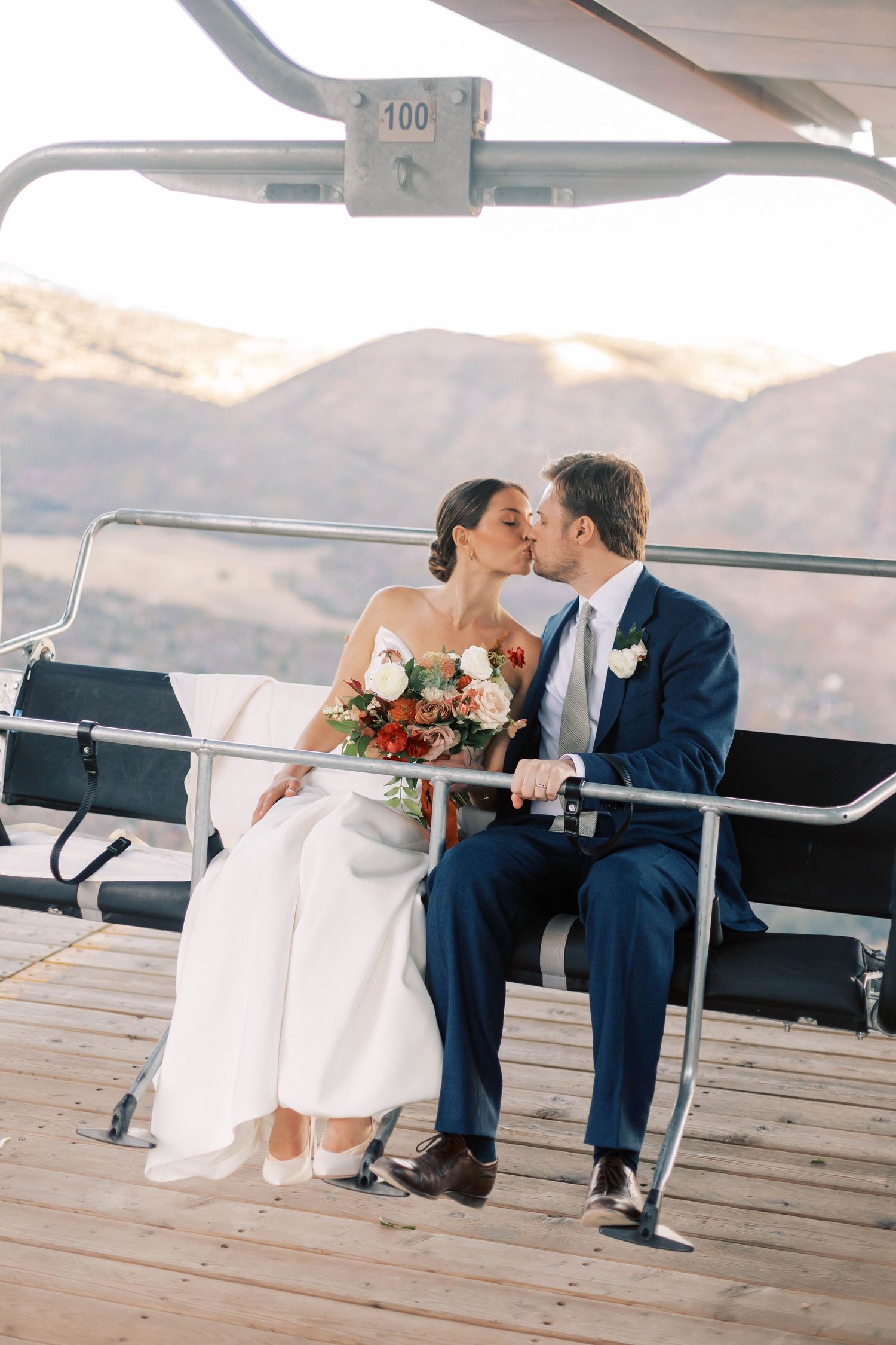 Happy couple sharing a kiss while seated on a chairlift with scenic mountain views.