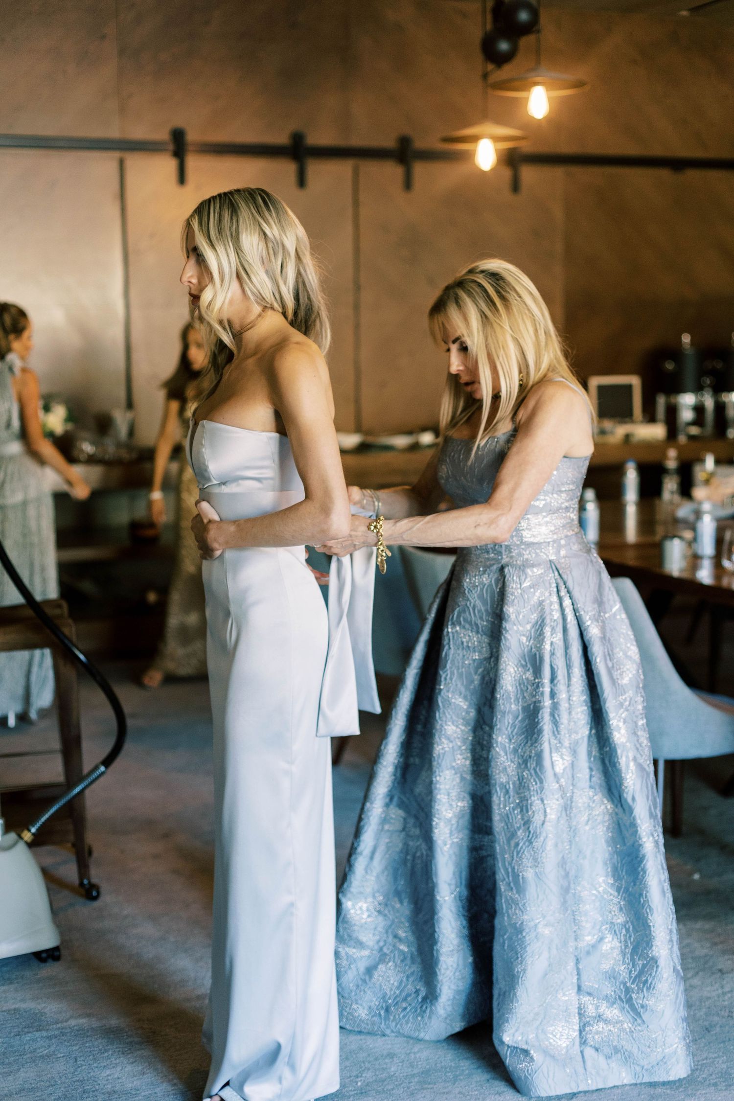 Wedding dress fitting scene with two women in formal attire in an industrial-style venue.