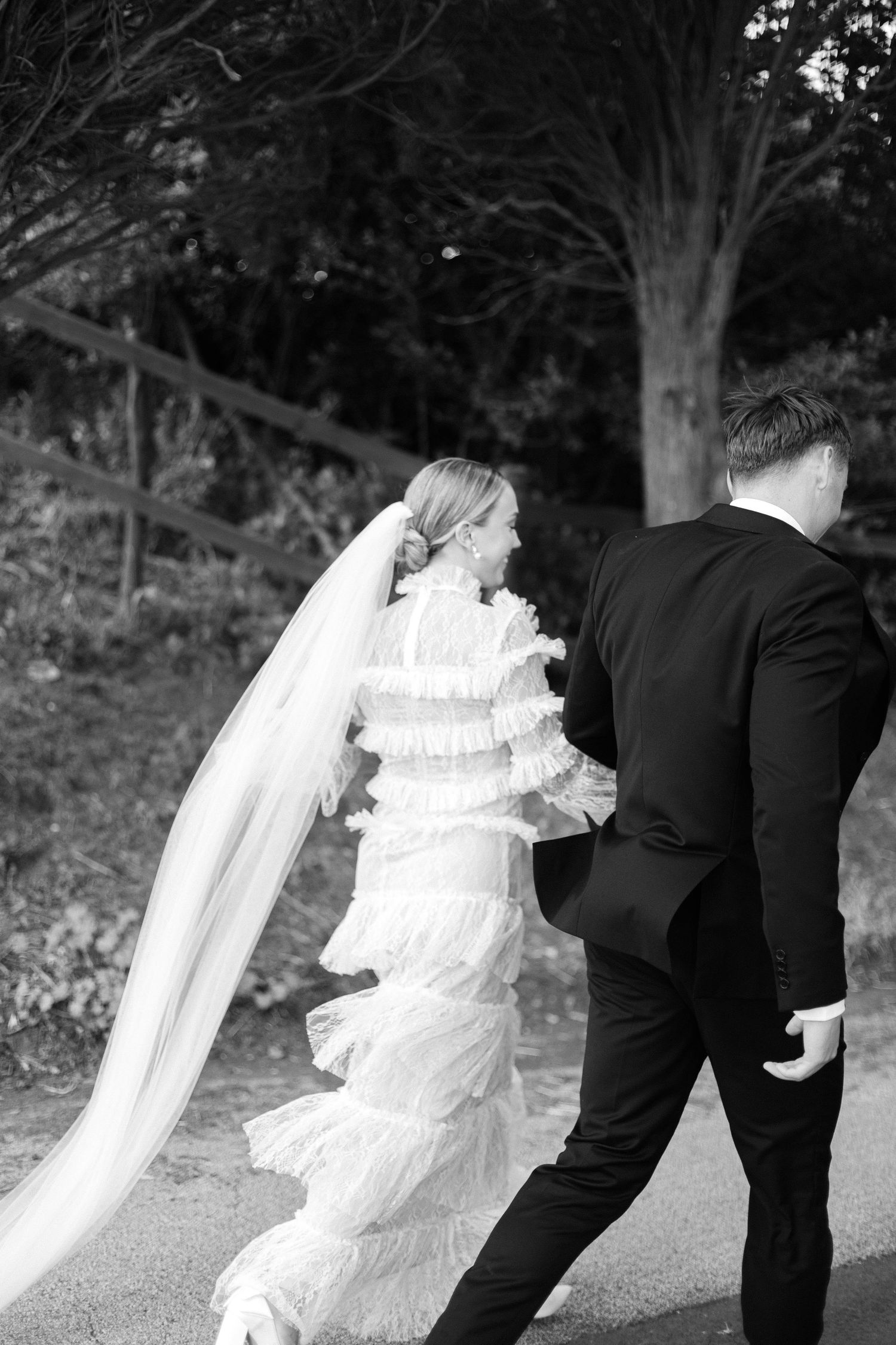 Black and white photo of newlyweds walking together on a path with a flowing white tiered dress and veil.