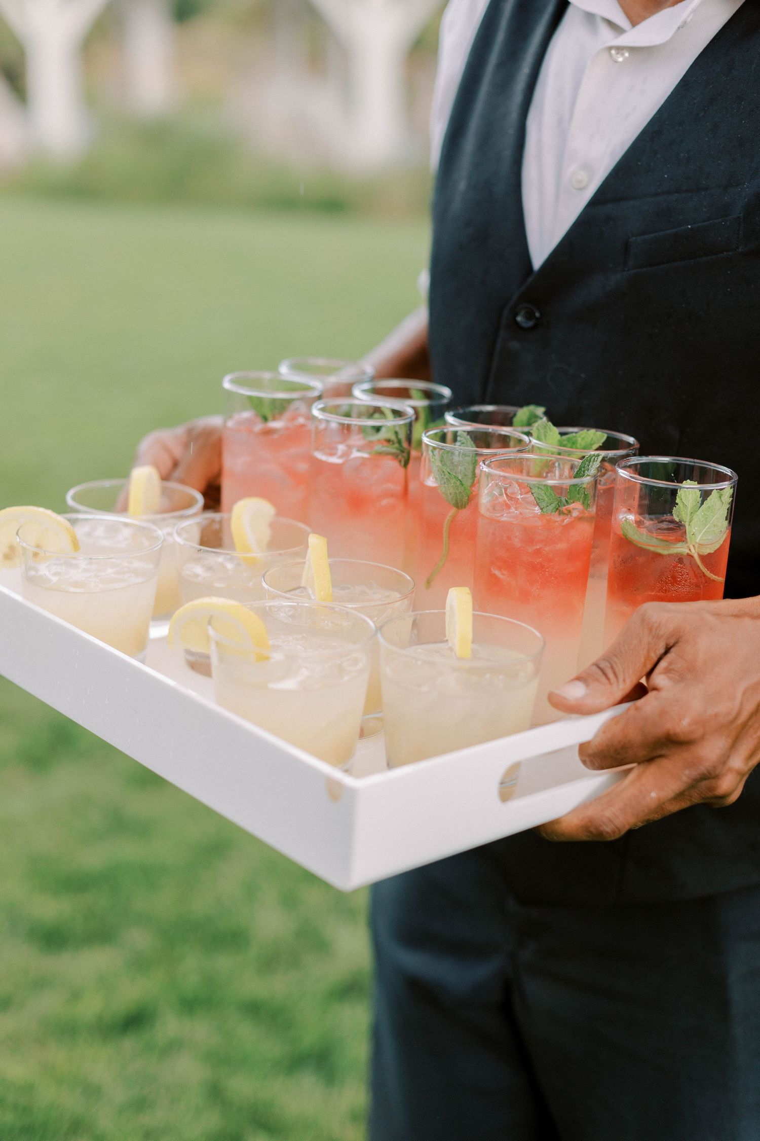 Server holding tray of pink and clear signature cocktails garnished with herbs.