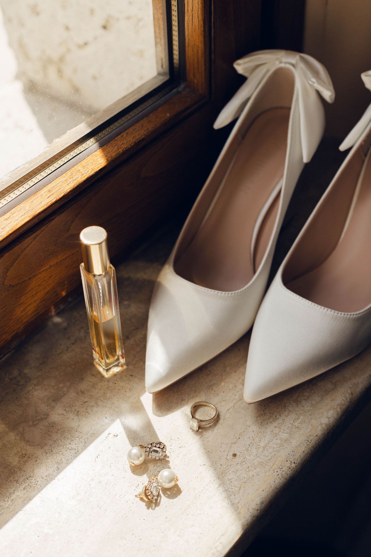White bridal heels and perfume bottle arranged near window with wedding jewelry.