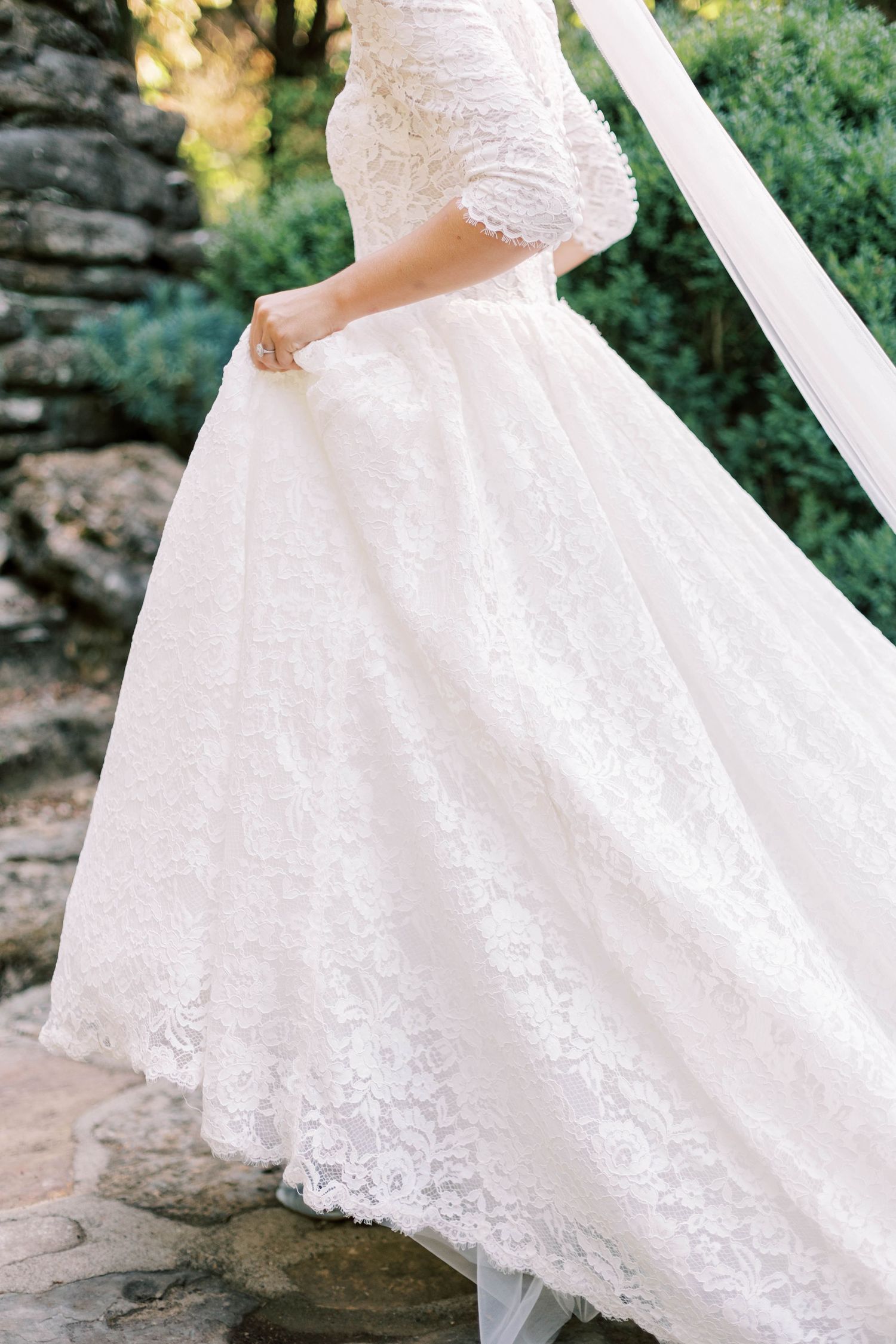 Close-up detail of a flowing white lace wedding dress with intricate embroidery on stone steps.